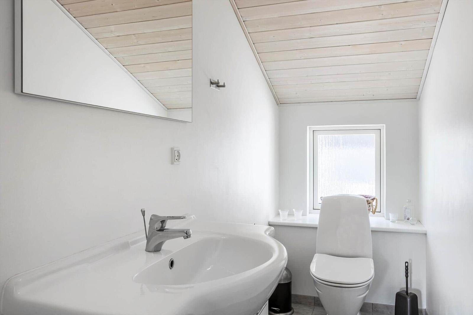 Badezimmer mit Waschbecken, Toilette und Fenster. Holzdecke und Spiegel sichtbar.
