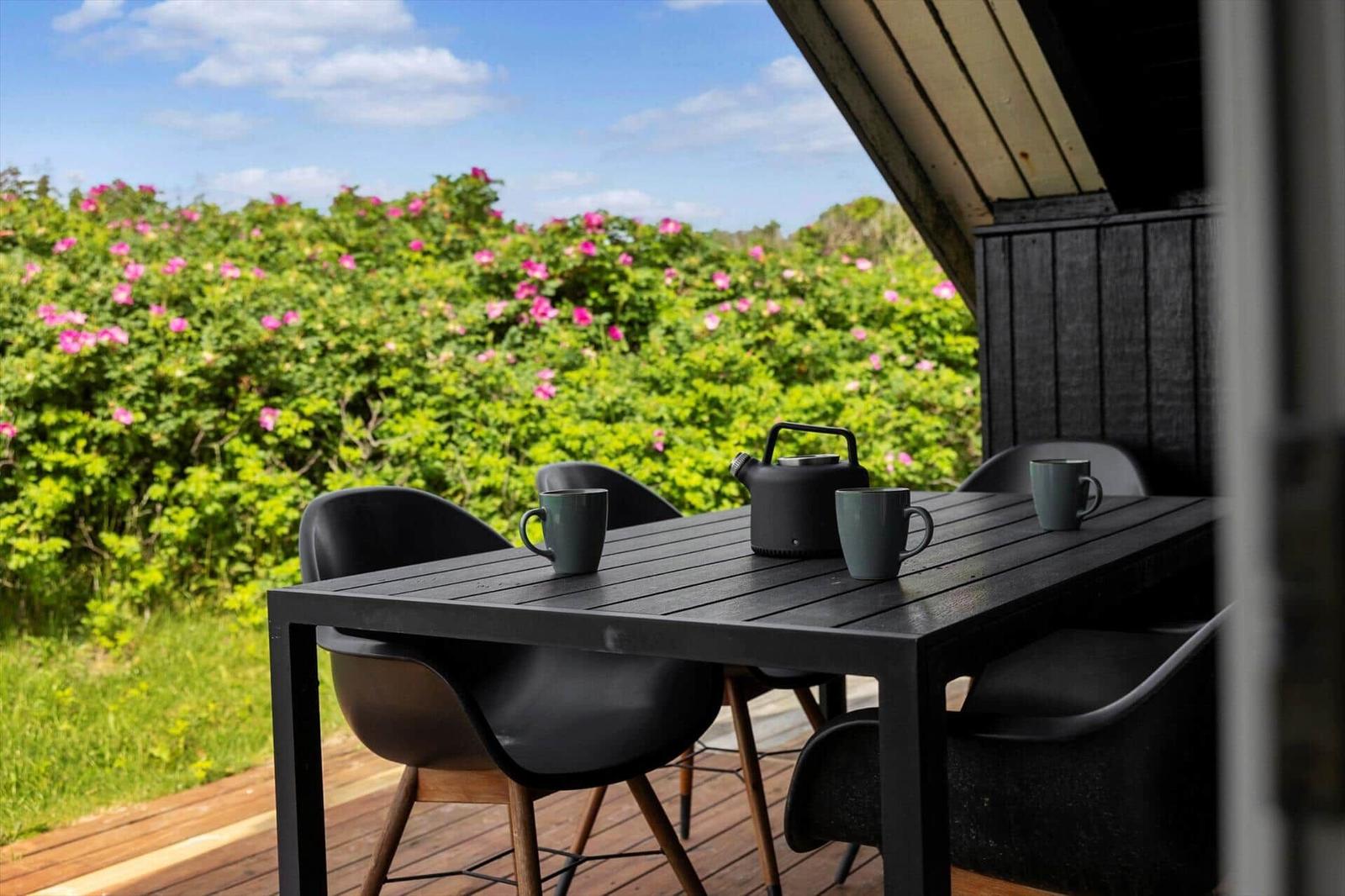 Terrasse mit Tisch, Stühlen und Teekanne vor blühenden Sträuchern.