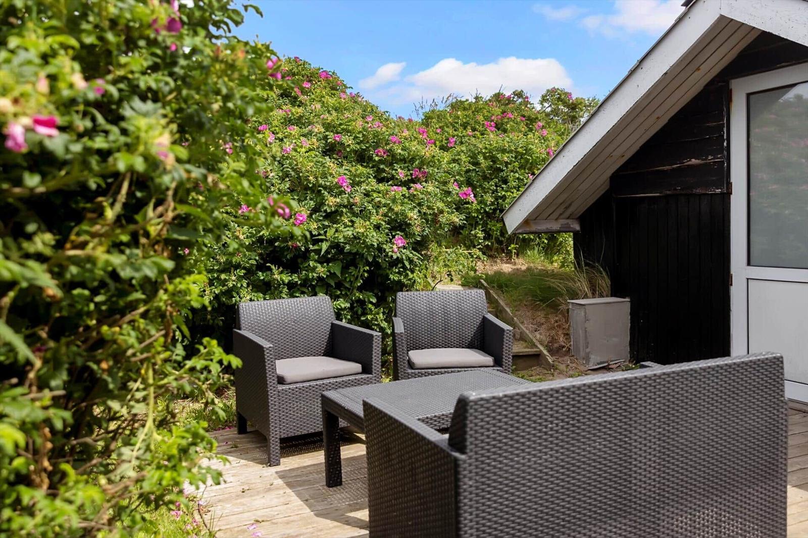 Terrasse mit drei Gartenstühlen und Tisch. Hintergrund: grüne Hecke mit rosa Blüten.