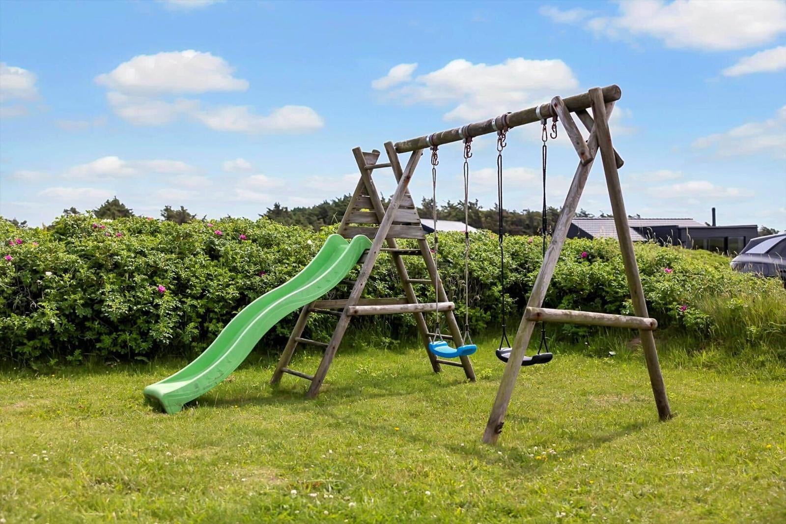 Holzschaukel mit grüner Rutsche im Garten. Hintergrund: grüne Hecke und blauer Himmel.