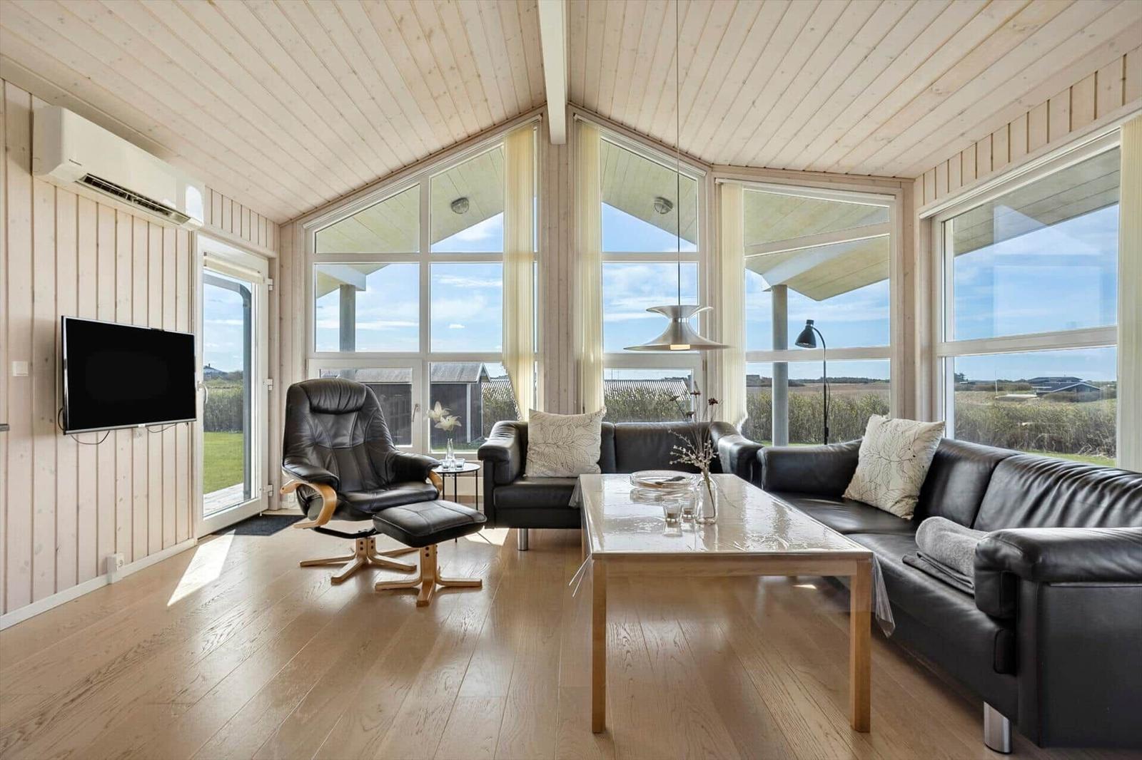 Wohnzimmer mit schwarzen Ledermöbeln, Holzboden und großen Fenstern mit Blick auf die Landschaft.