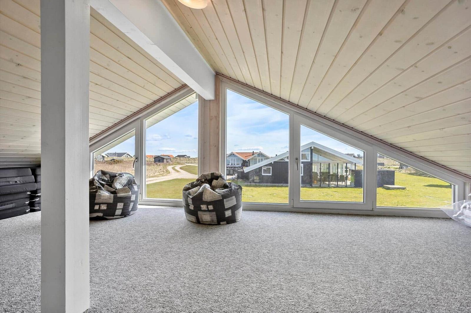 Raum mit großen Fenstern, grauem Teppich und zwei Sitzkissen. Blick auf Grünflächen und Häuser.