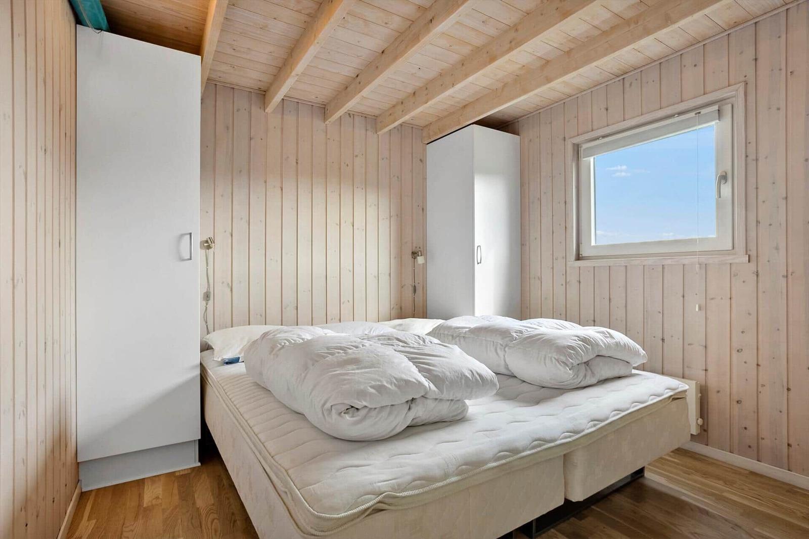 Schlafzimmer mit Holzwandverkleidung, weißem Bett und Fenster mit Blick auf den Himmel.