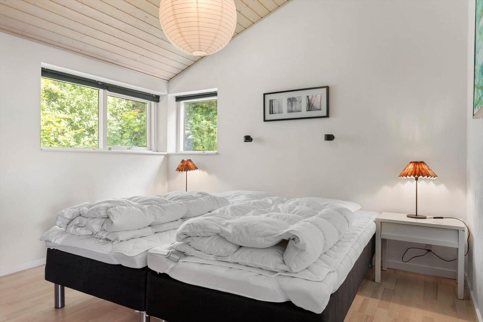 Schlafzimmer mit Doppelbett, zwei Fenstern und Nachttisch mit Lampe.