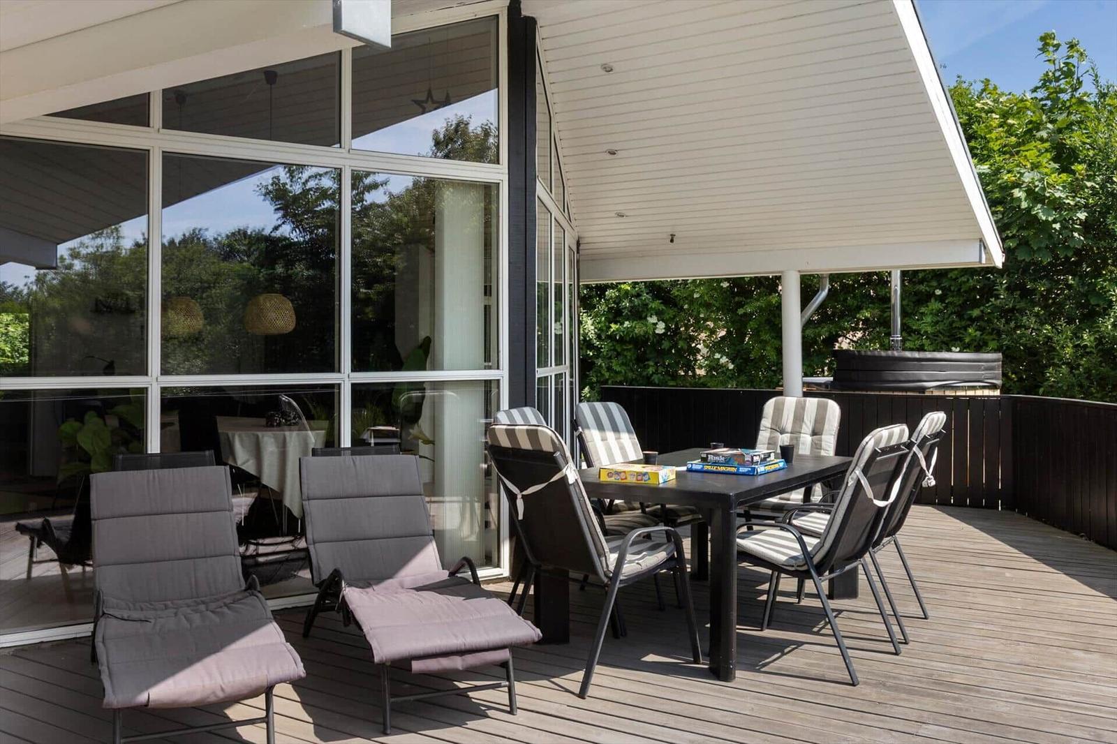 Terrasse mit Tisch, Stühlen und Liegestühlen. Hintergrund: Glasfassade und grüne Bäume.