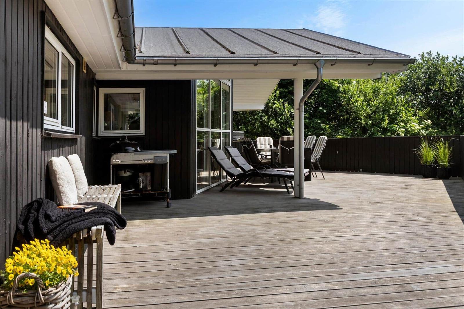 Terrasse mit Liegestühlen, Grill und Pflanzen. Holzdecke und Sicht auf Grün.