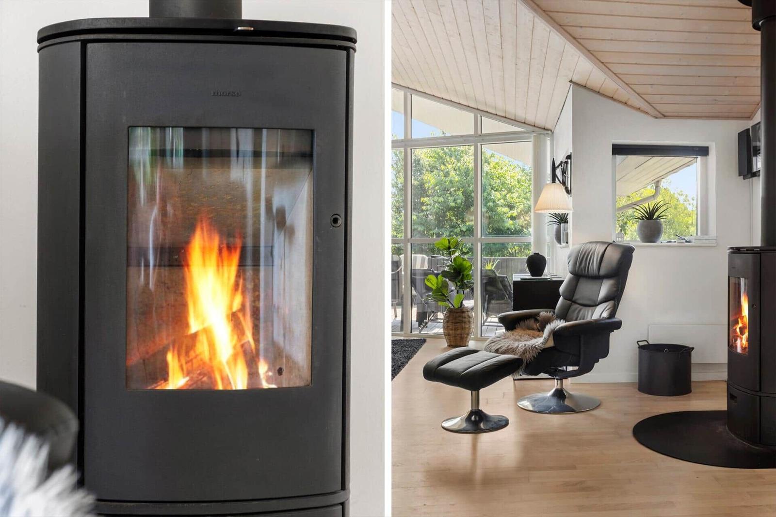 Ein Kamin mit brennendem Holz steht in einem Raum mit Sessel und großem Fenster.