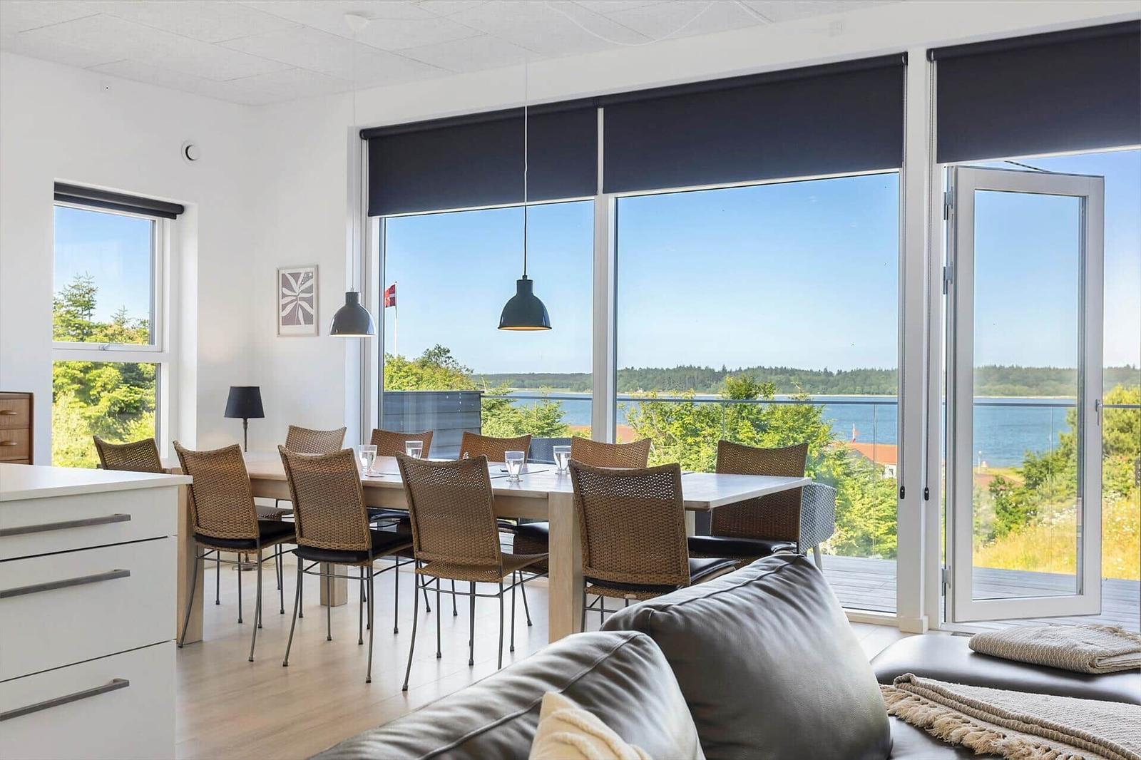 Essbereich mit Blick auf Wasser und Wald. Tisch, Stühle und große Fenster.