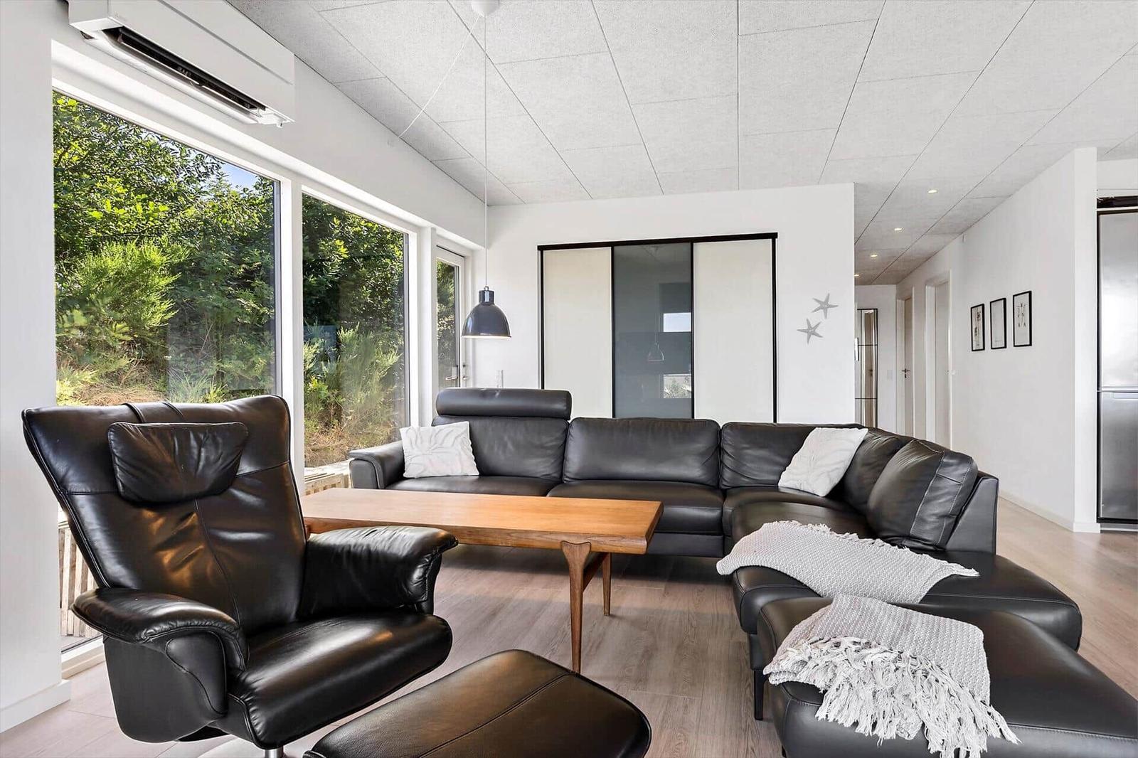 Wohnzimmer mit schwarzer Ledercouch, Holztisch und großem Fensterblick auf Grün.