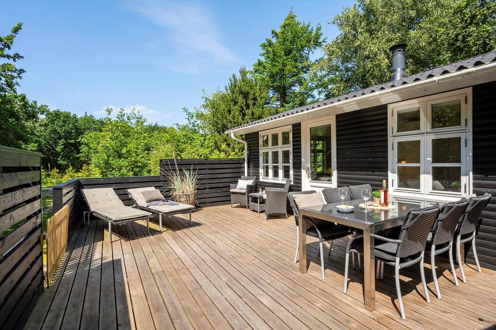 Terrasse mit Loungemöbeln und Esstisch. Holzterrasse, schwarze Fassade, grüne Umgebung.