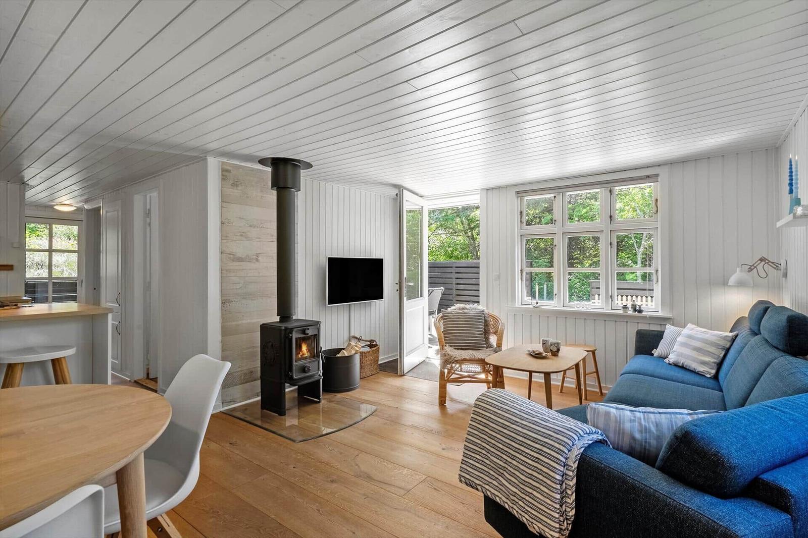 Wohnraum mit Kamin, Sofa und Küchenbereich. Holzboden und weiße Wände.