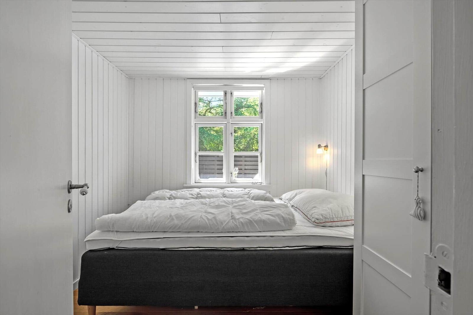 Schlafzimmer mit weißem Holz und Bett. Fenster zeigt grüne Bäume.