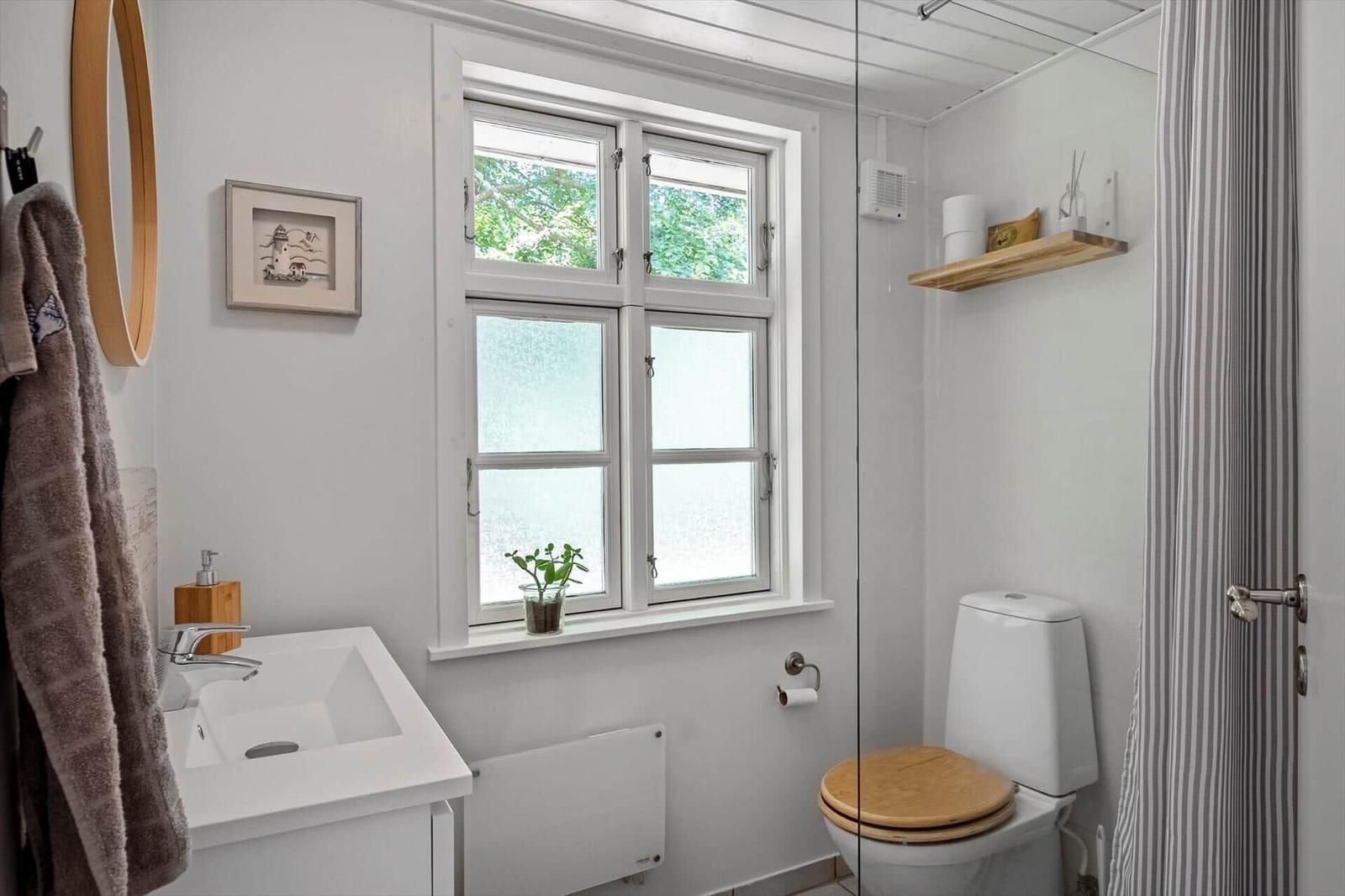Badezimmer mit Fenster, Toilette und Waschbecken. Holztoilettenkappe und Wandregal.