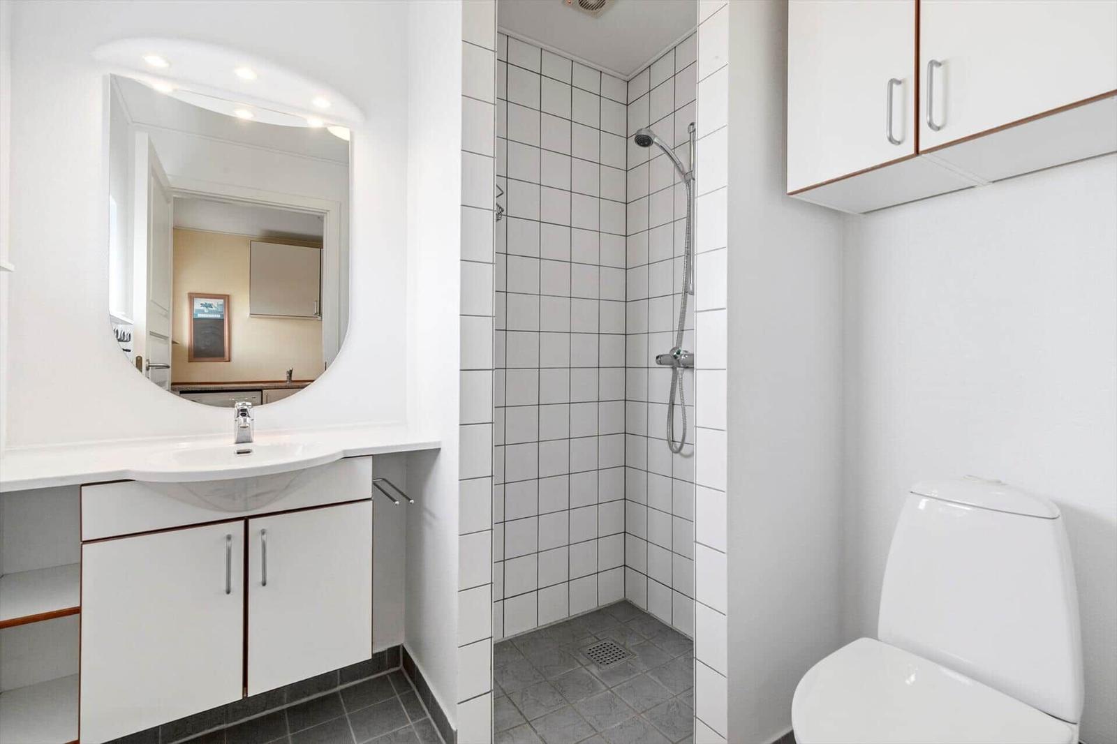 Badezimmer mit Dusche, Waschbecken und Toilette. Weiße Fliesen und Schränke.