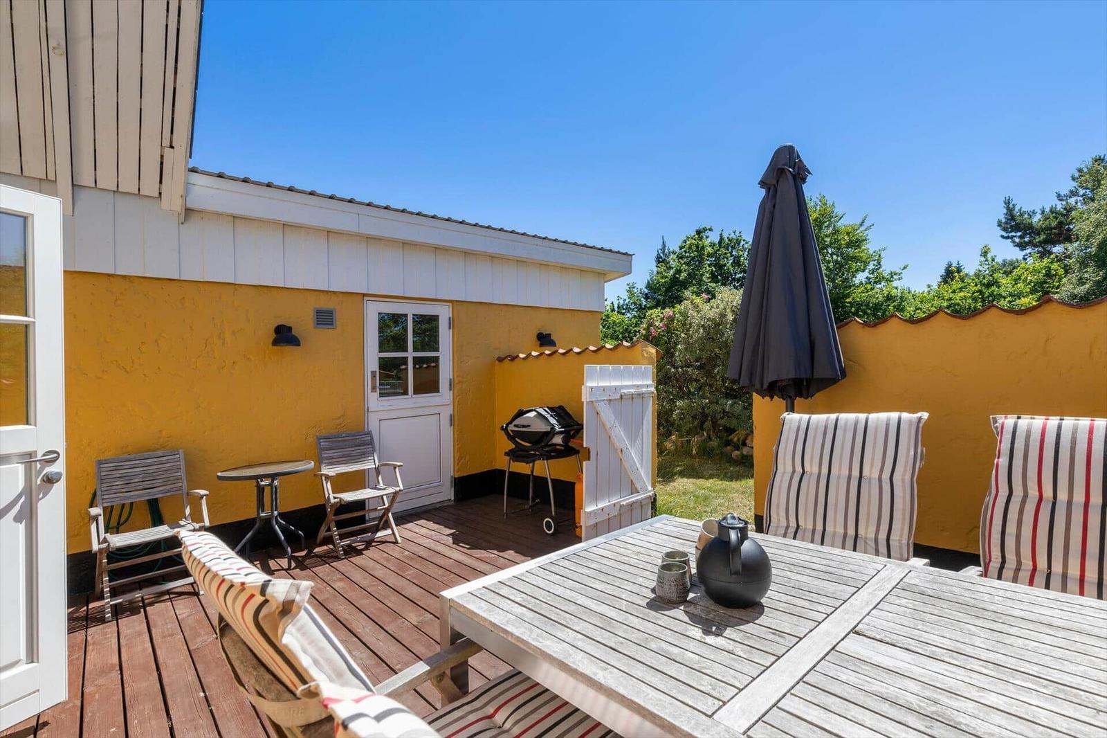 Terrasse mit Holztisch, Stühlen und Grill. Gelber Hauswänden und geschlossener Sonnenschirm.