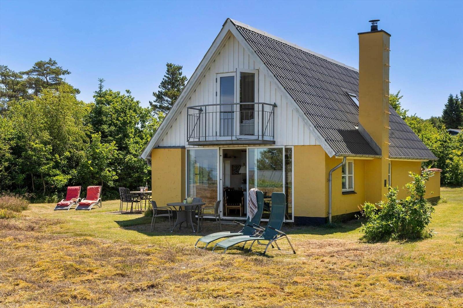 Gelbes Haus mit Balkon und Gartenmöbeln. Terrasse mit Tisch und Stühlen. Hintergrund: Bäume.