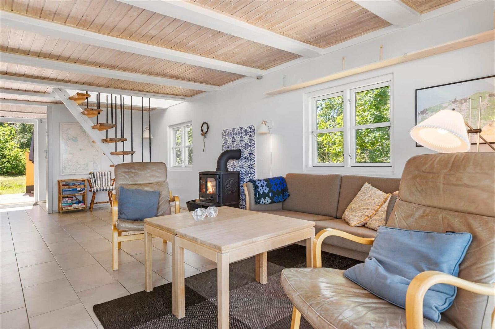 Wohnzimmer mit Sofa, zwei Sesseln, Tisch und Kaminofen. Holzdecke und Fenster mit Blick auf Grün.