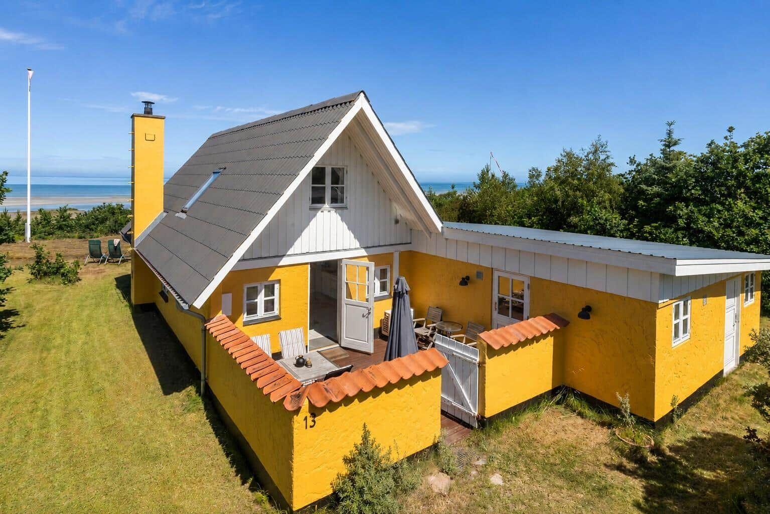Ein gelbes Haus mit Terrasse und Meerblick. Weiße Fenster und grauer Dachfirst.