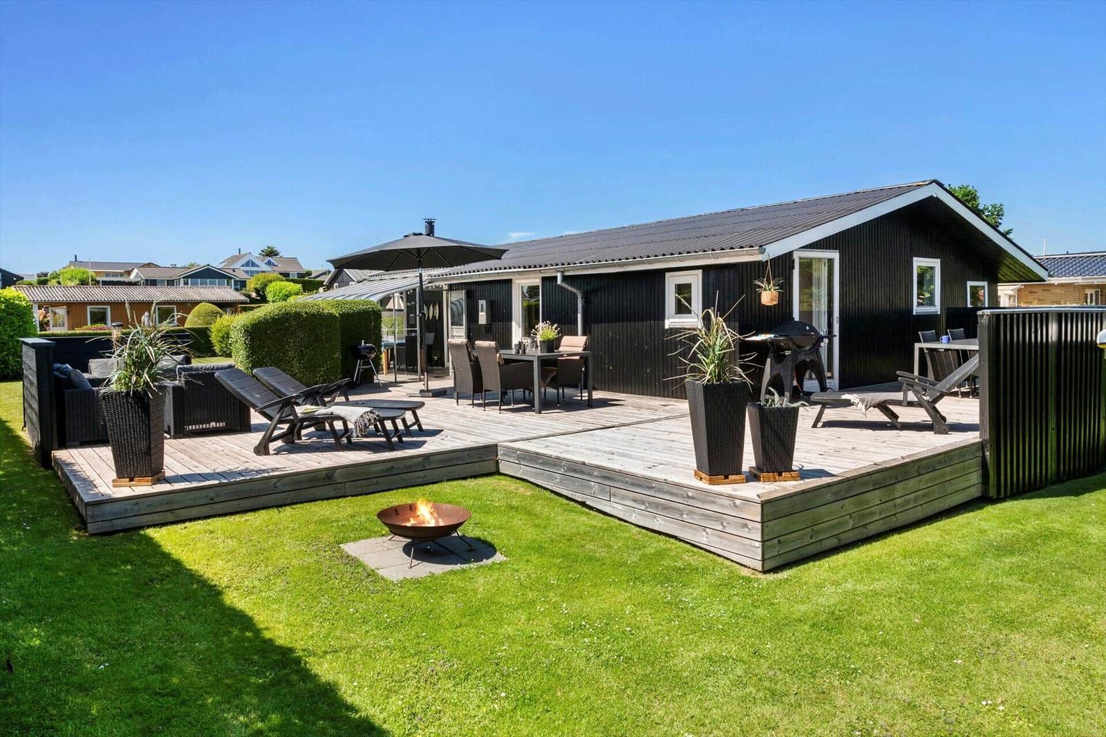 A holiday home with a large terrace, garden, and fire pit. Furniture and plants are arranged on the terrace.