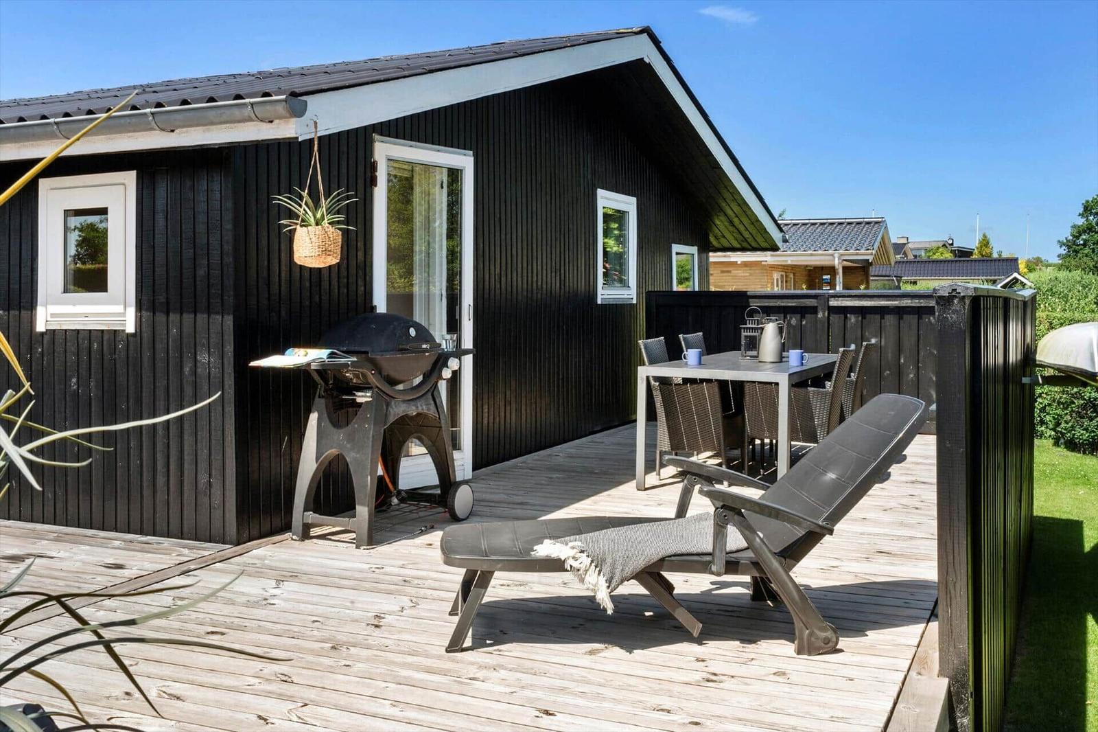 Deck with grill, table, chairs, and lounge chair in front of black wood house.