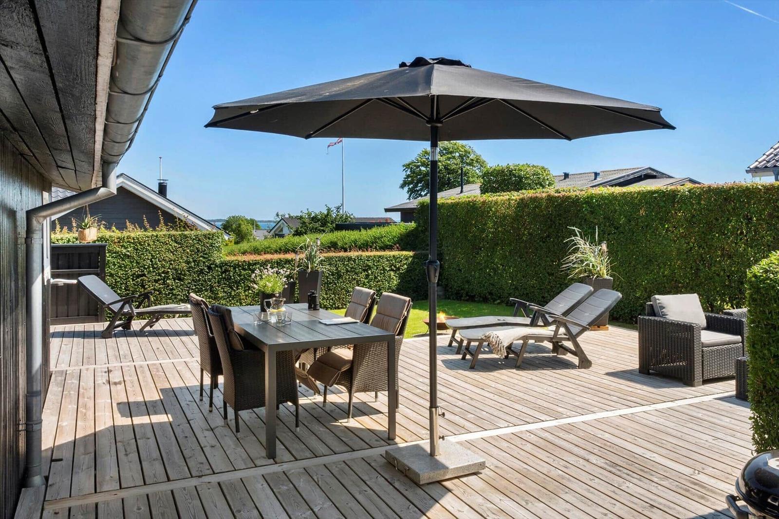 Deck with table, chairs, and umbrella. Background: green hedge and houses.