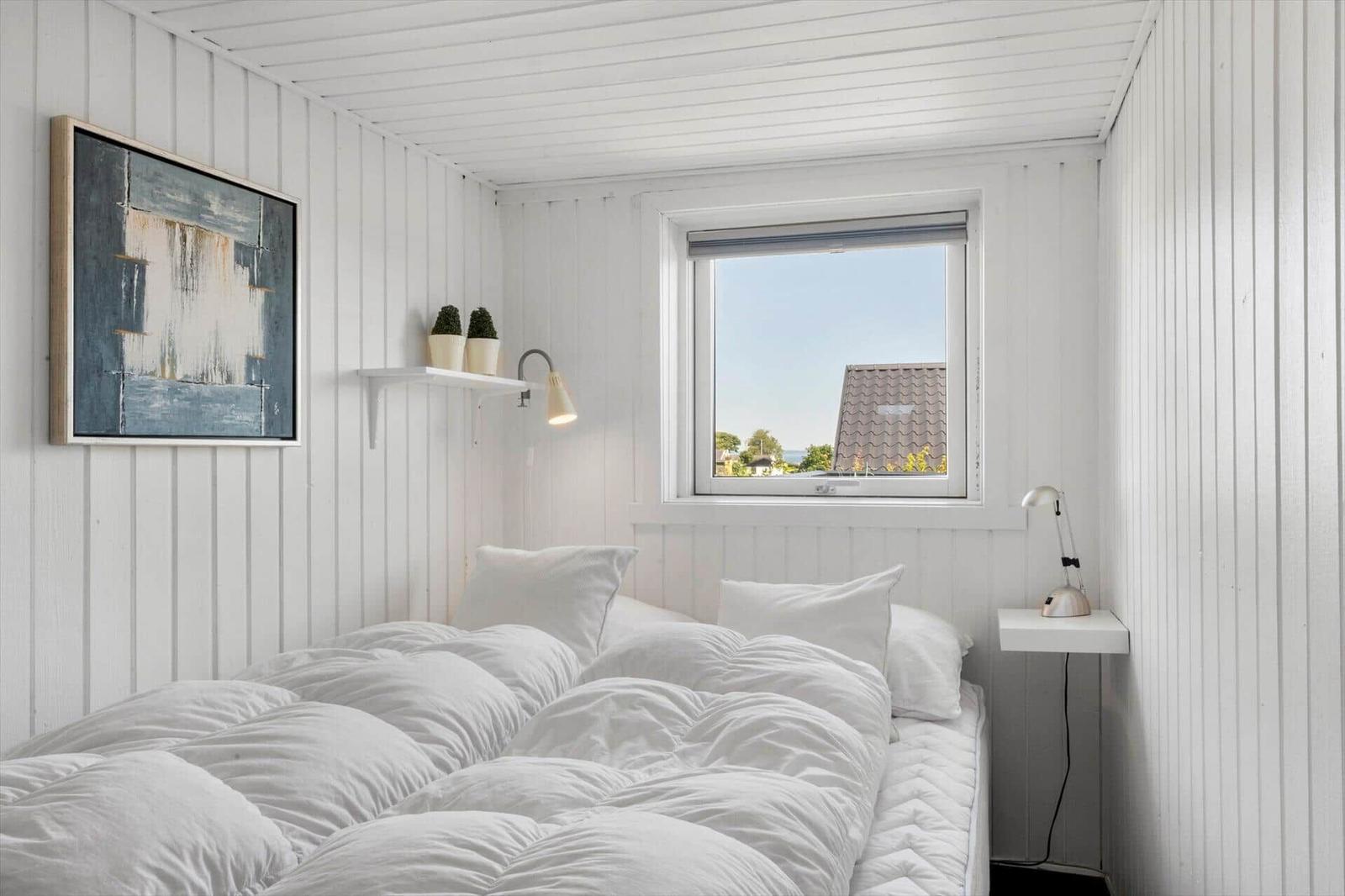 A bedroom with a white bed, wall art, and window view of a roof.