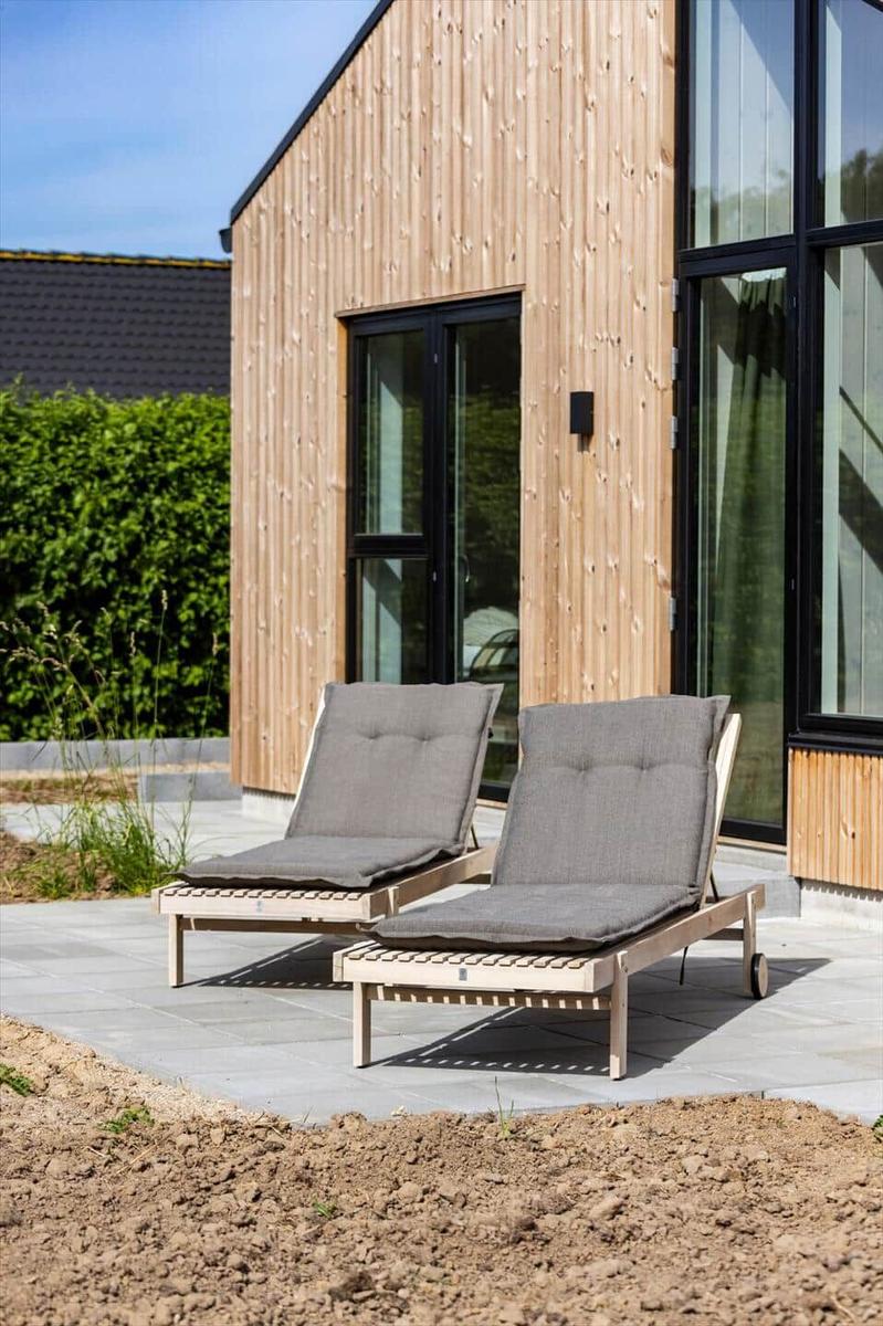 Zwei Liegestühle mit Kissen stehen auf einer Terrasse vor einem Holzhaus mit großen Fenstern.