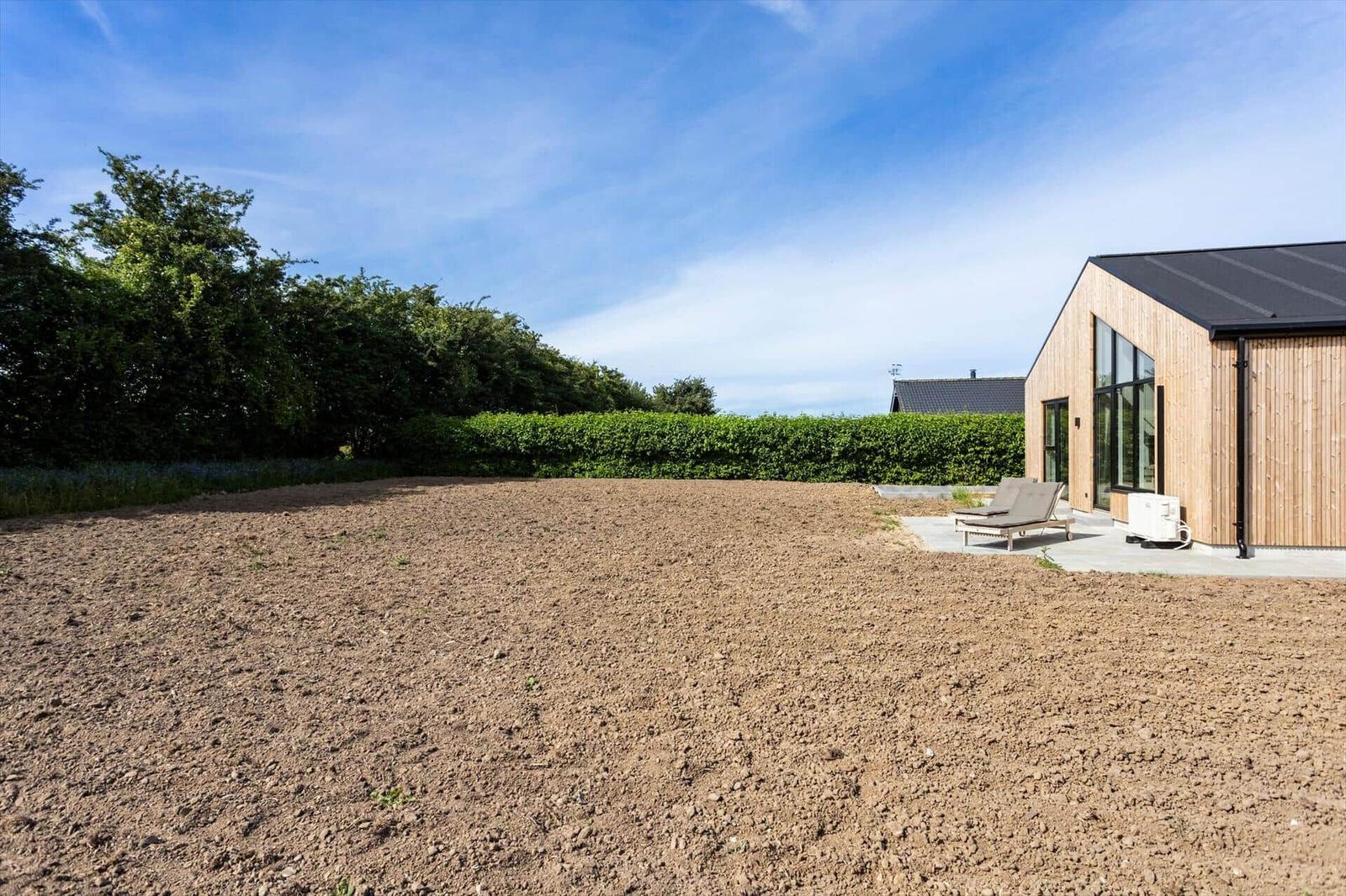 Moderne Holzhütte mit Terrasse und Liegen im Garten. Hintergrund: Hecke und blauer Himmel.