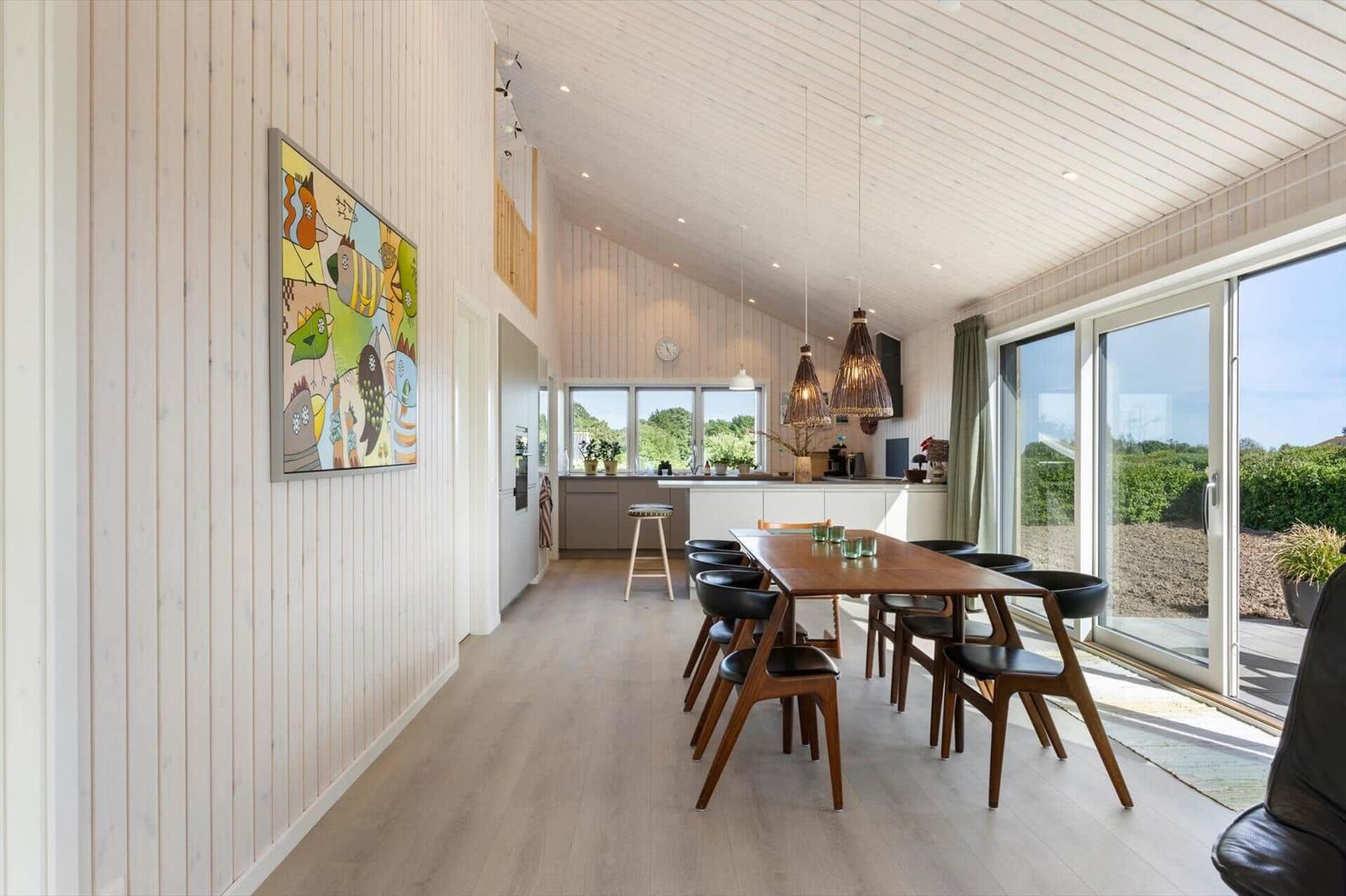 Essbereich mit Holztisch, Stühlen und Küchenbereich. Große Fenster und Wandbild.