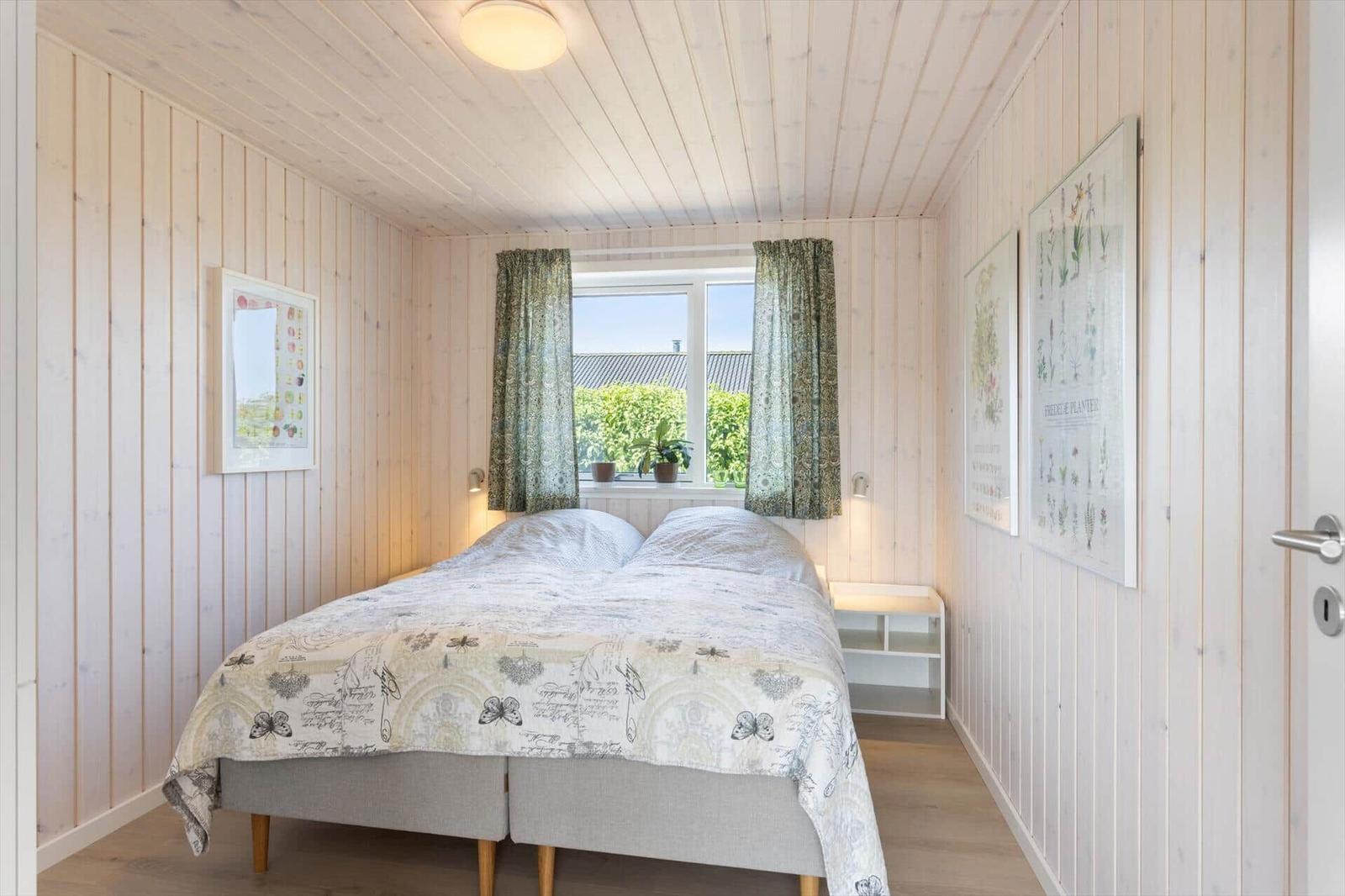 Schlafzimmer mit Bett, Fenster und Holzwänden. An den Wänden hängen Bilder.