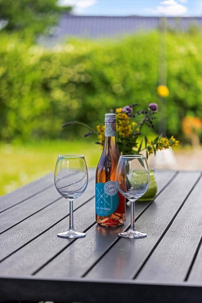 Auf einem Tisch stehen zwei Weingläser und eine Flasche alkoholfreies Rosé. Im Hintergrund Blumen und Grün.