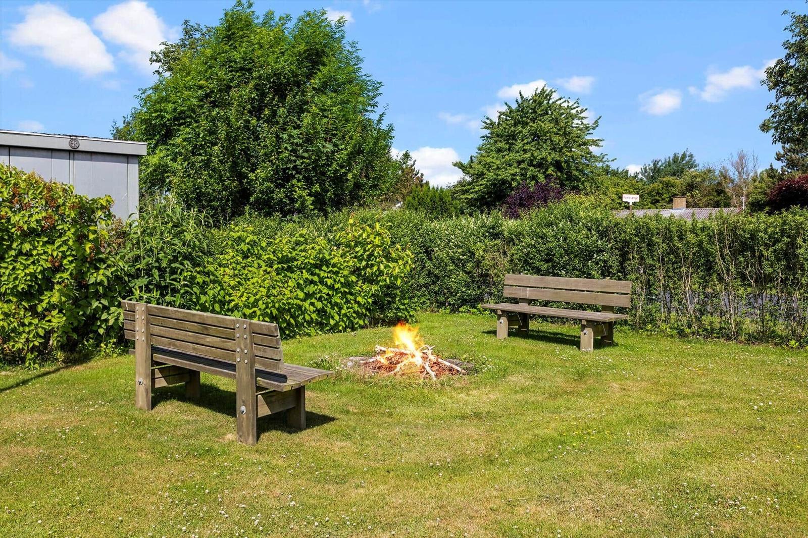 Garten mit Holzbank, Feuerstelle und grüner Hecke unter blauem Himmel.