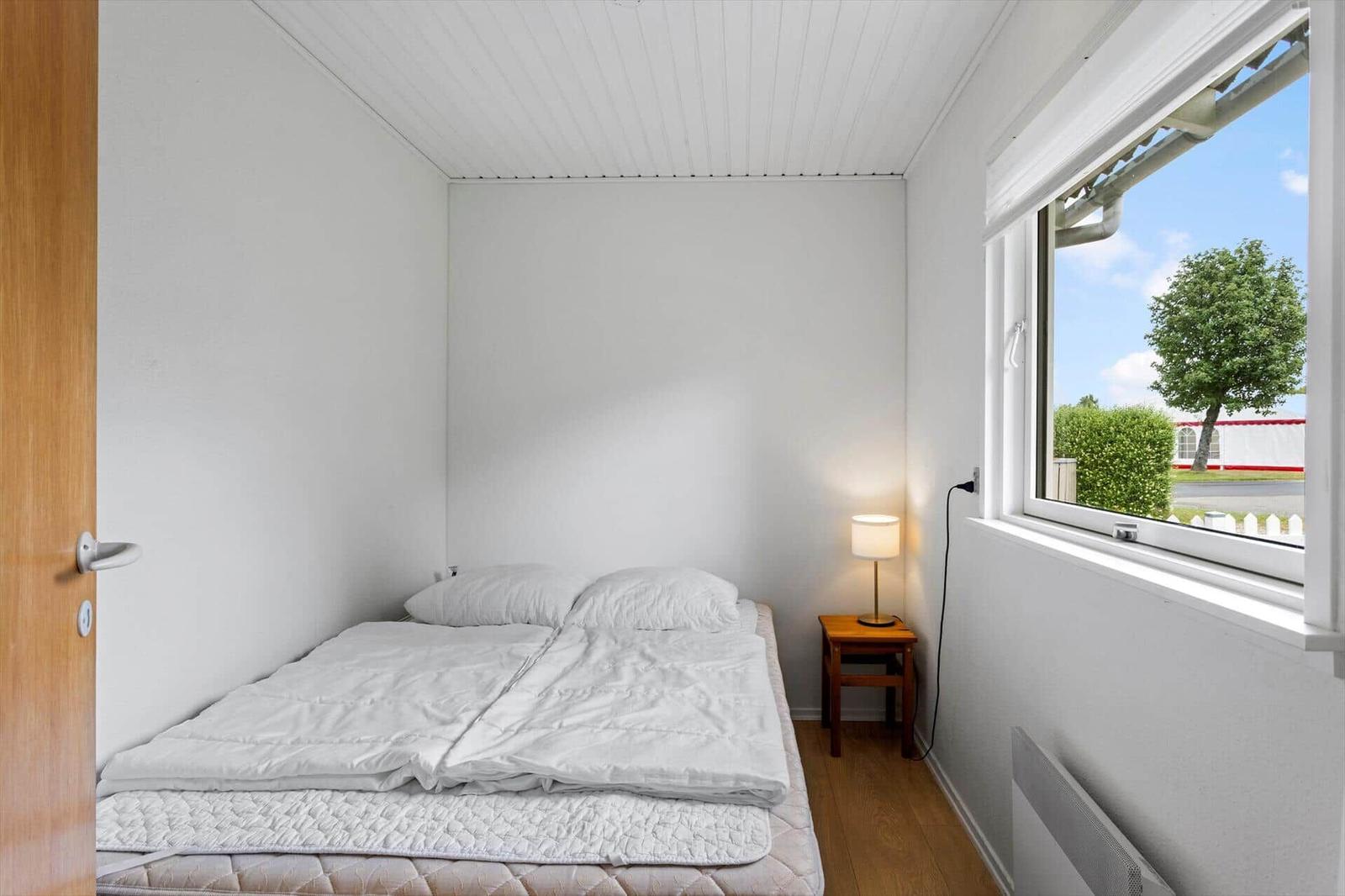 Ein Schlafzimmer mit Bett, Nachttisch und Lampe. Durch das Fenster ist ein Baum sichtbar.