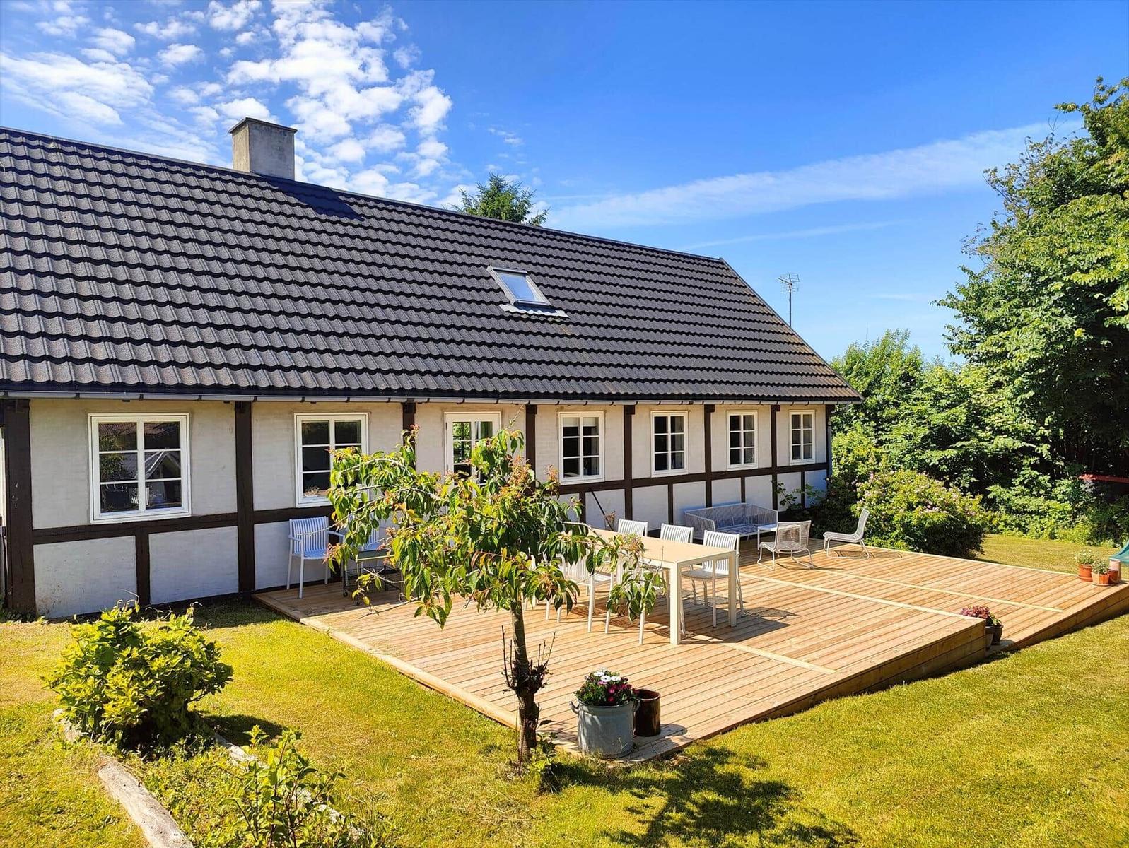 A house with a terrace, garden, and a tree. There is a table set and chairs.