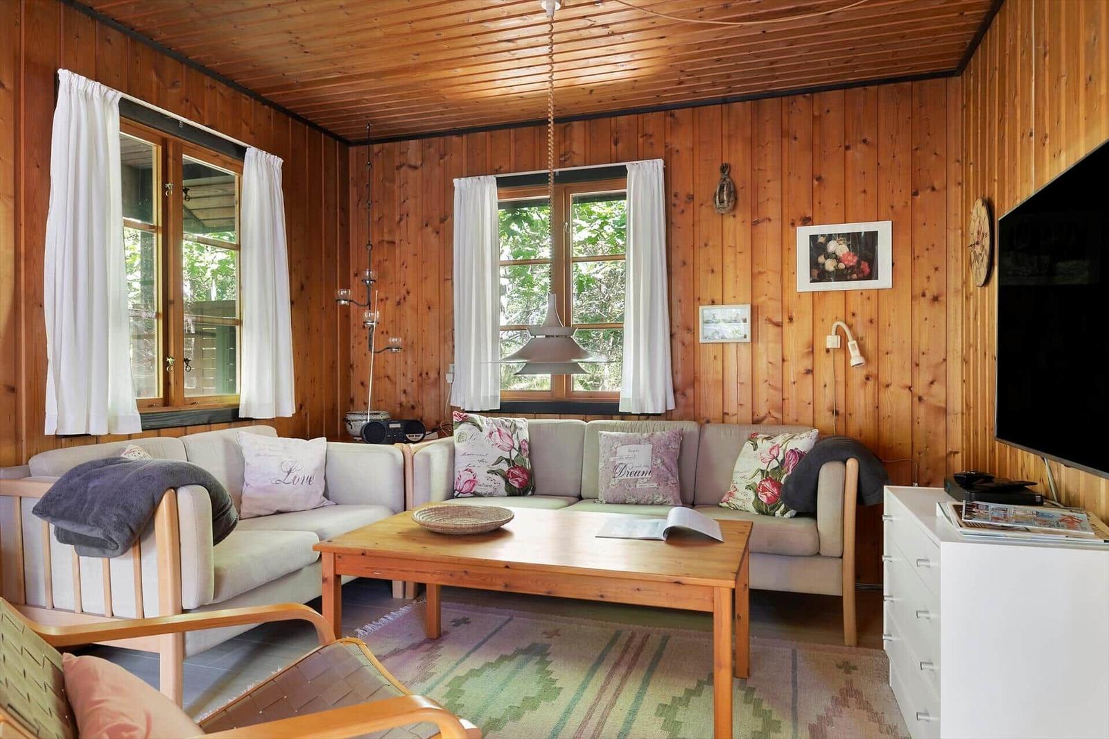 Wohnzimmer mit Holzwänden, Sofa, Couchtisch und Fenstern mit weißen Vorhängen.