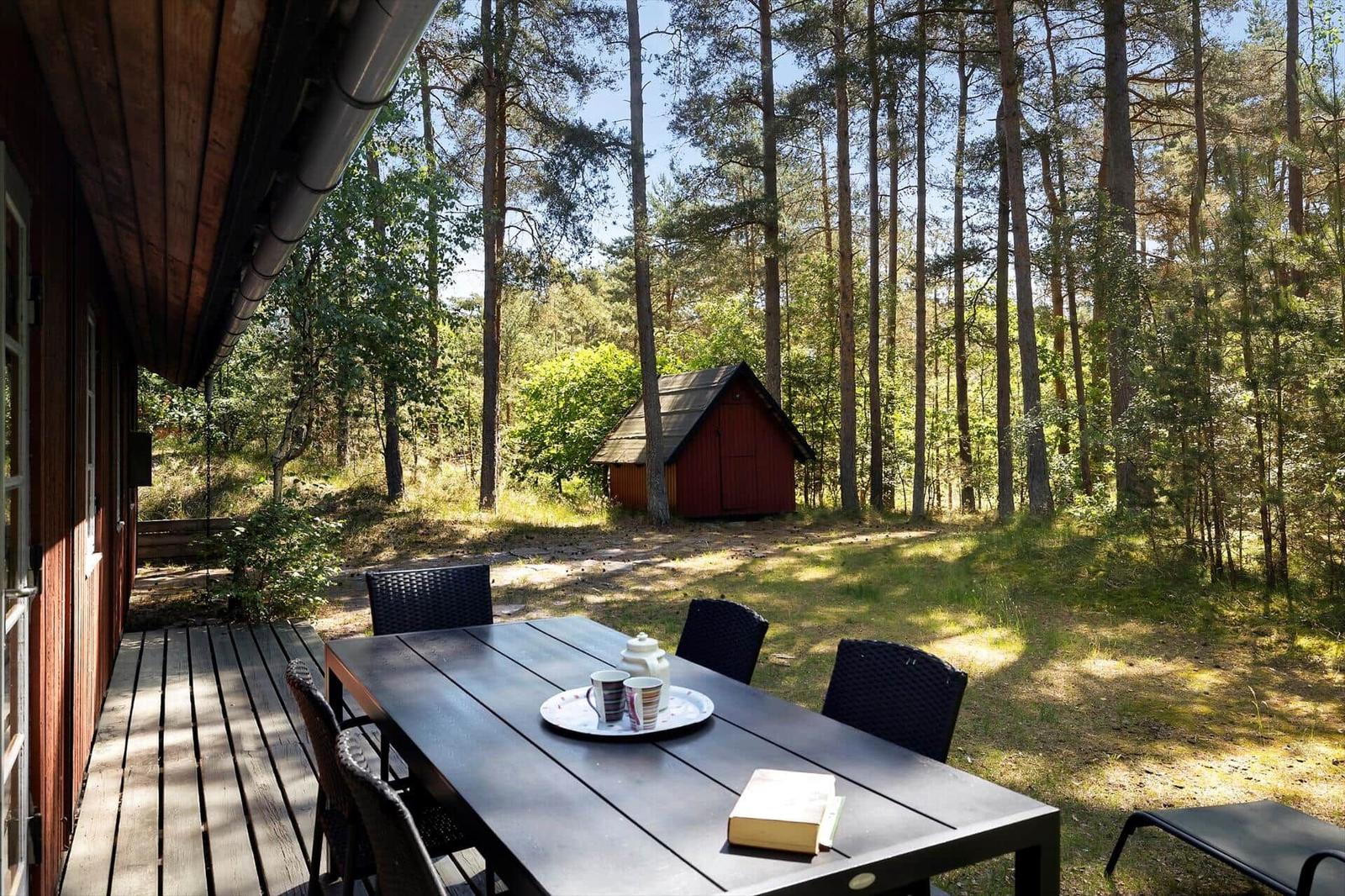 Terrasse mit Tisch und Stühlen im Wald. Im Hintergrund steht eine rote Hütte.