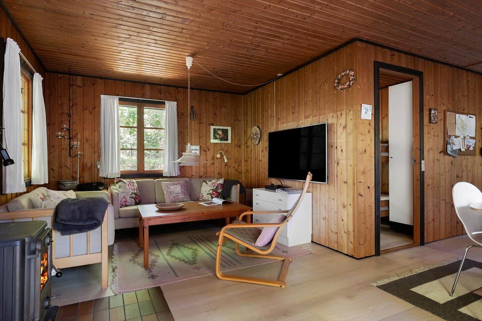 Wohnzimmer mit Holzwand, Sofa, Fernseher und Kaminofen. Durchgang zu Schlafzimmer.