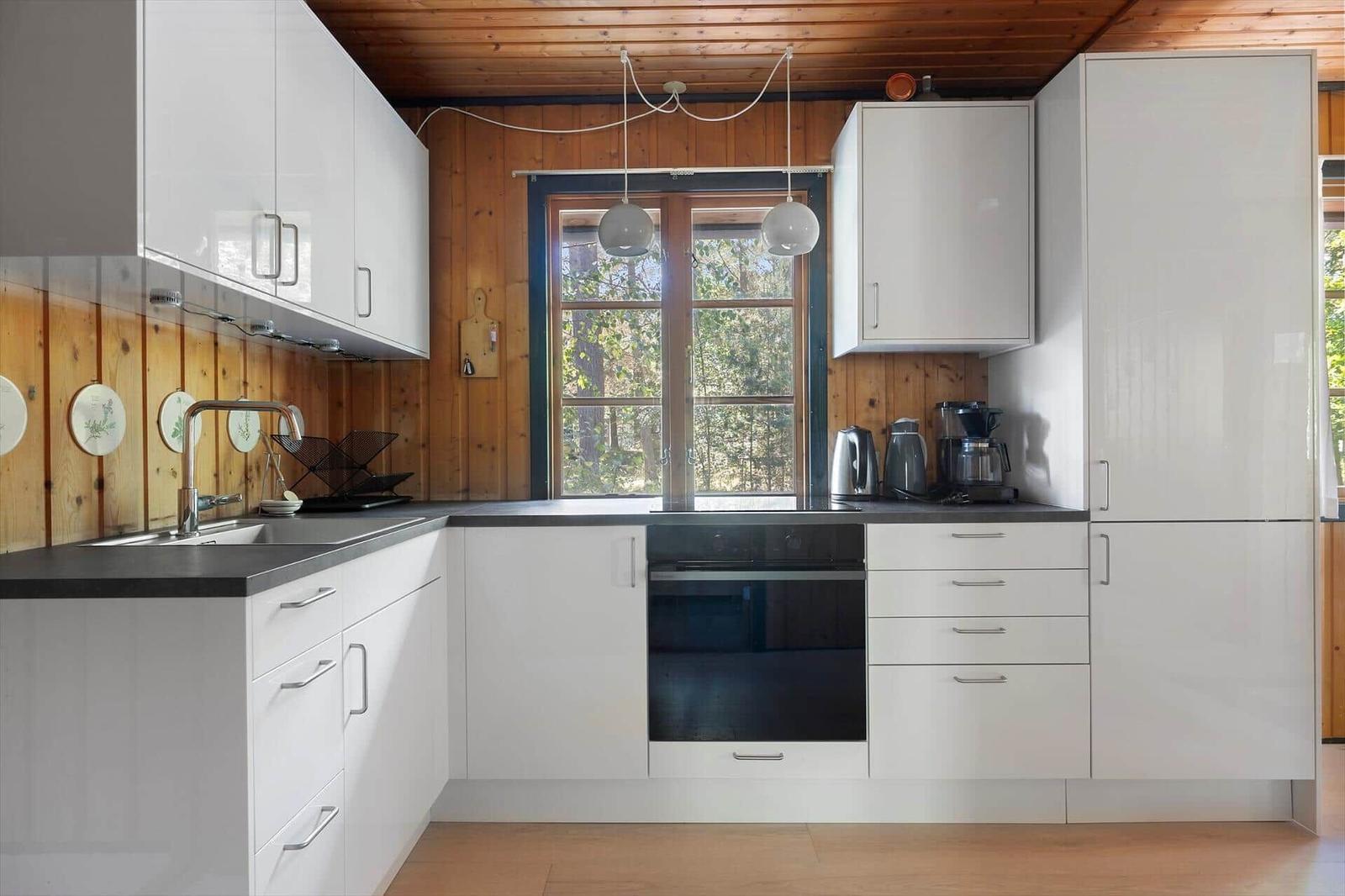 Küche mit weißen Schränken, dunkler Arbeitsplatte und Holzwand. Fenster mit Blick auf Wald.