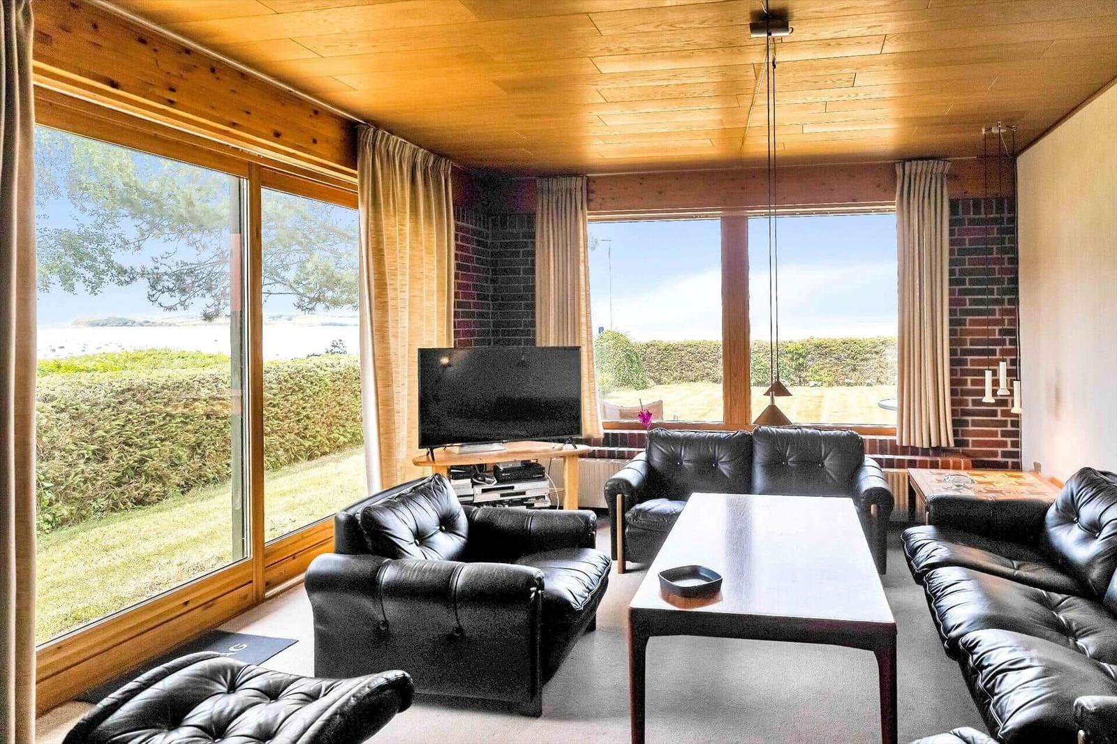 Living room with black leather furniture, wooden ceiling, and large windows overlooking green areas.