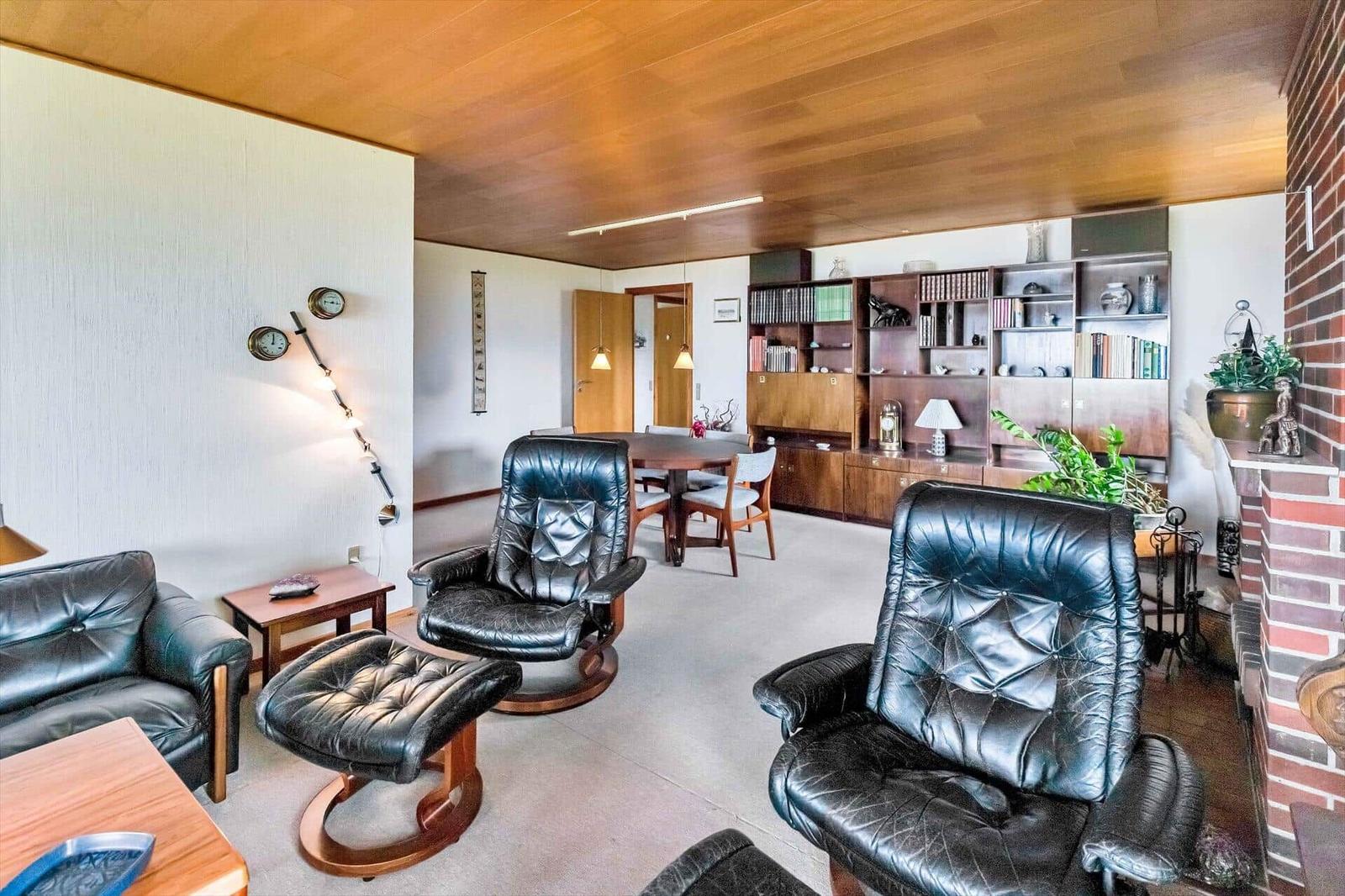 Living and dining area with leather sofa, chairs, and bookcase. Wooden ceiling and fireplace visible.