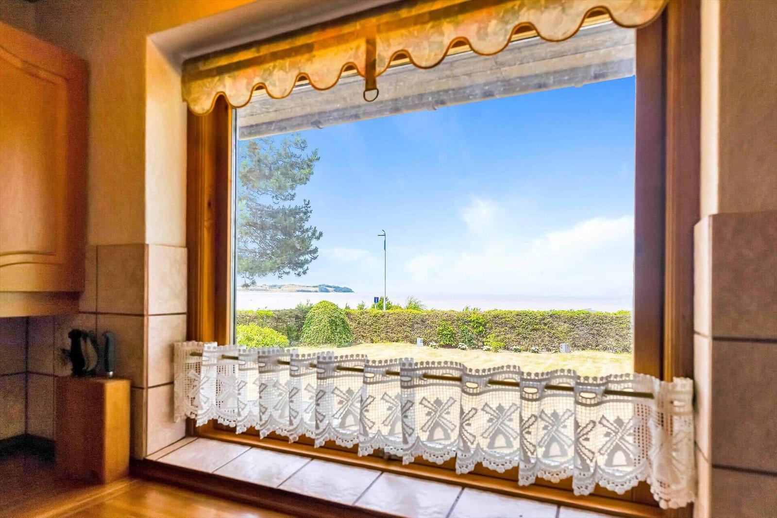 Kitchen window with sea and green view, white lace curtain and wooden frame.
