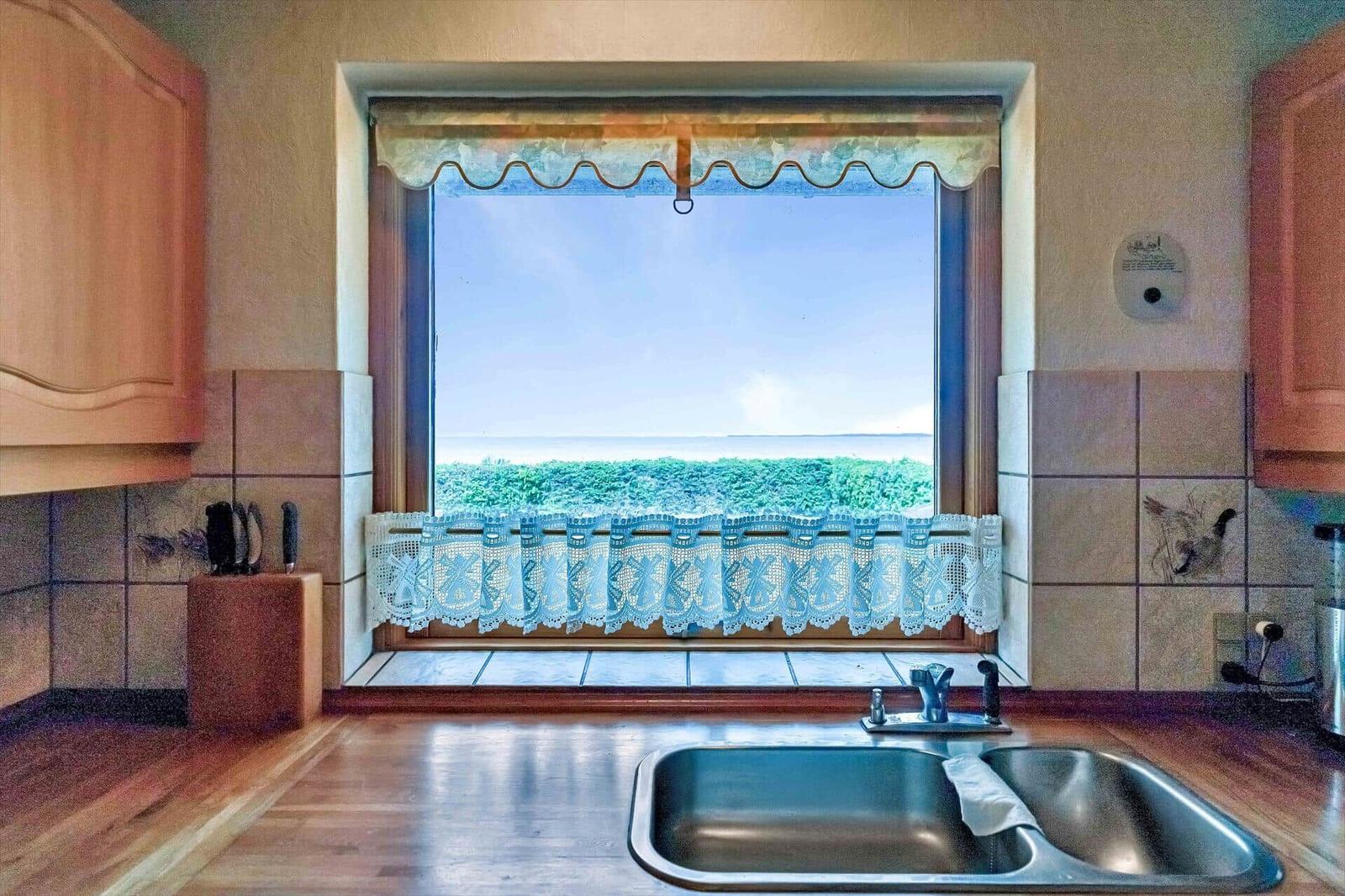 Kitchen with view of green field and sky. Stainless steel sink, wooden countertop, and window with curtain.