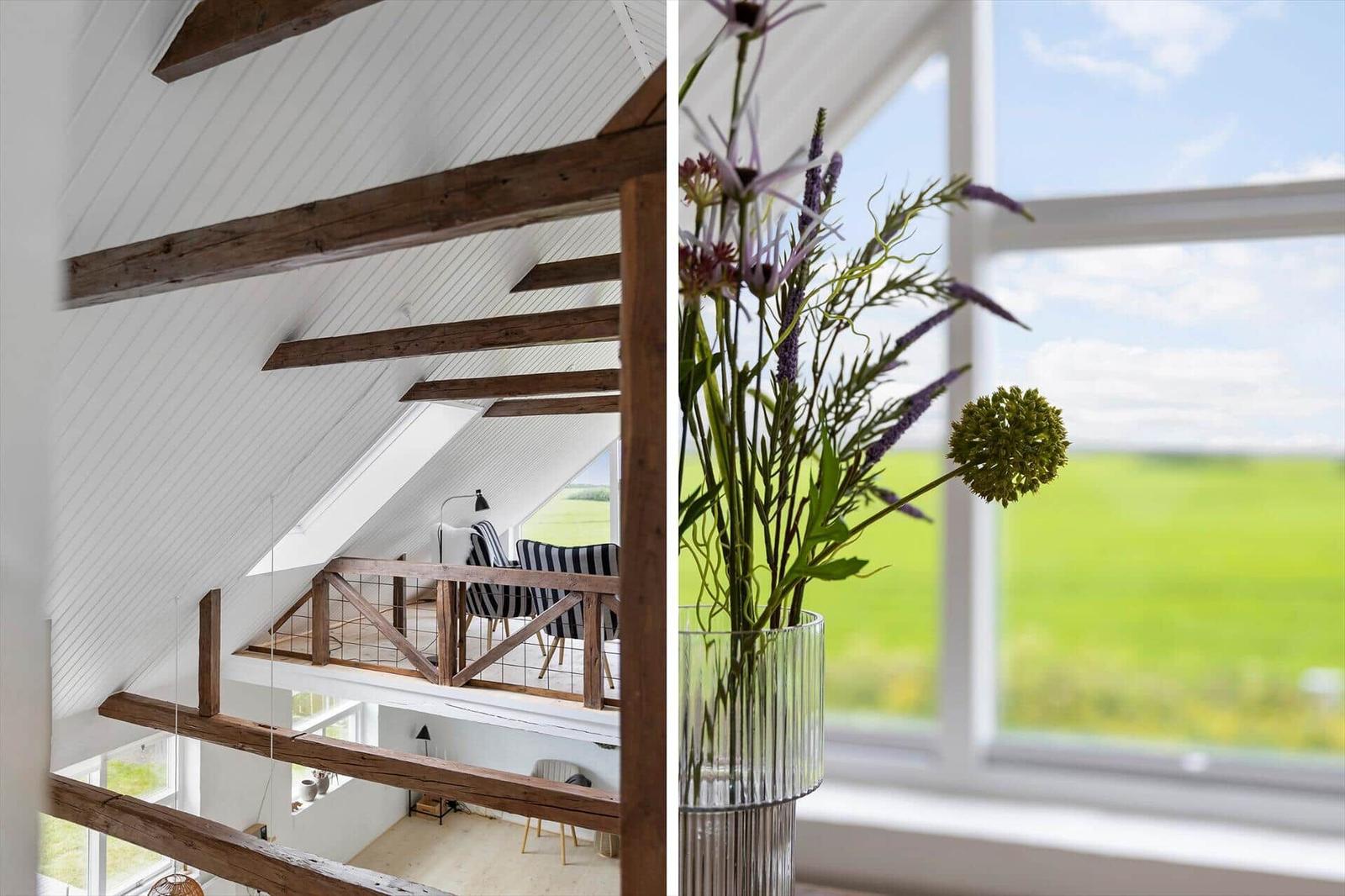Dachgeschoss mit Holzbalken und Aussicht auf grünes Feld. Blumen in Glasvase vor Fenster.