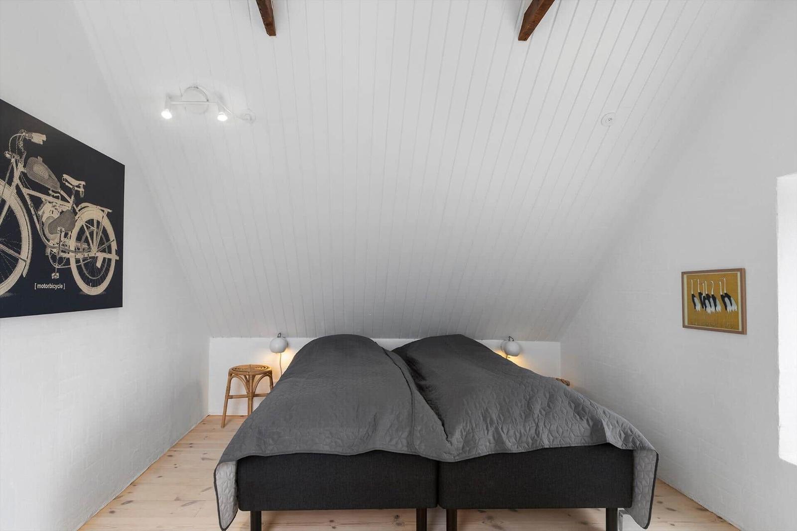 Schlafzimmer mit Doppelbett, Holzboden und schrägen weißen Wänden. Wandbilder und Nachttische sind vorhanden.