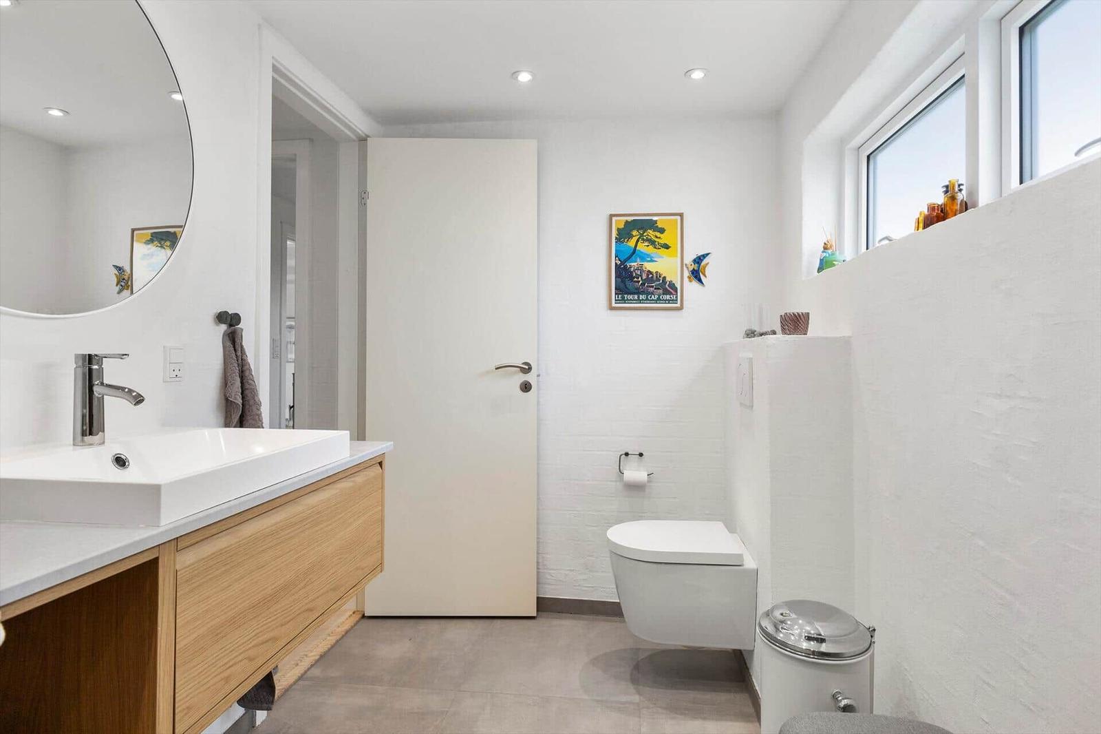 Badezimmer mit Waschtisch, Toilette und Fenster. Wandbild und runder Spiegel sichtbar.
