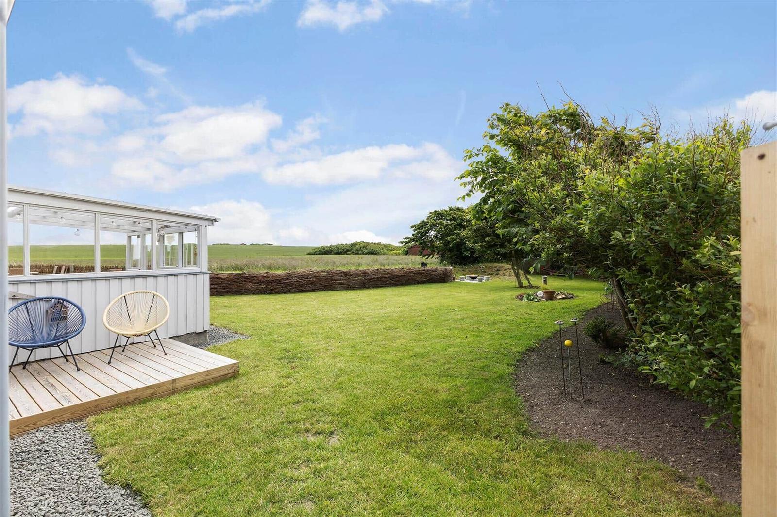 Hinterhof mit Grasfläche, Terrasse und zwei Stühlen. Im Hintergrund Landschaft mit Bäumen.