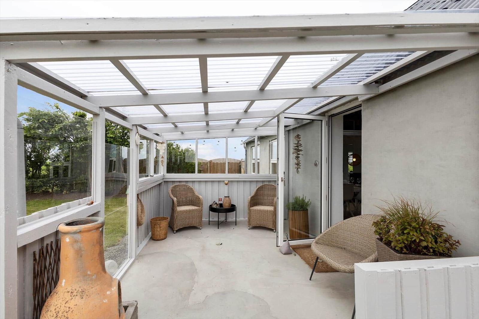 Terrasse mit Glasdach, zwei Stühlen und Pflanzen. Durchgang ins Haus.
