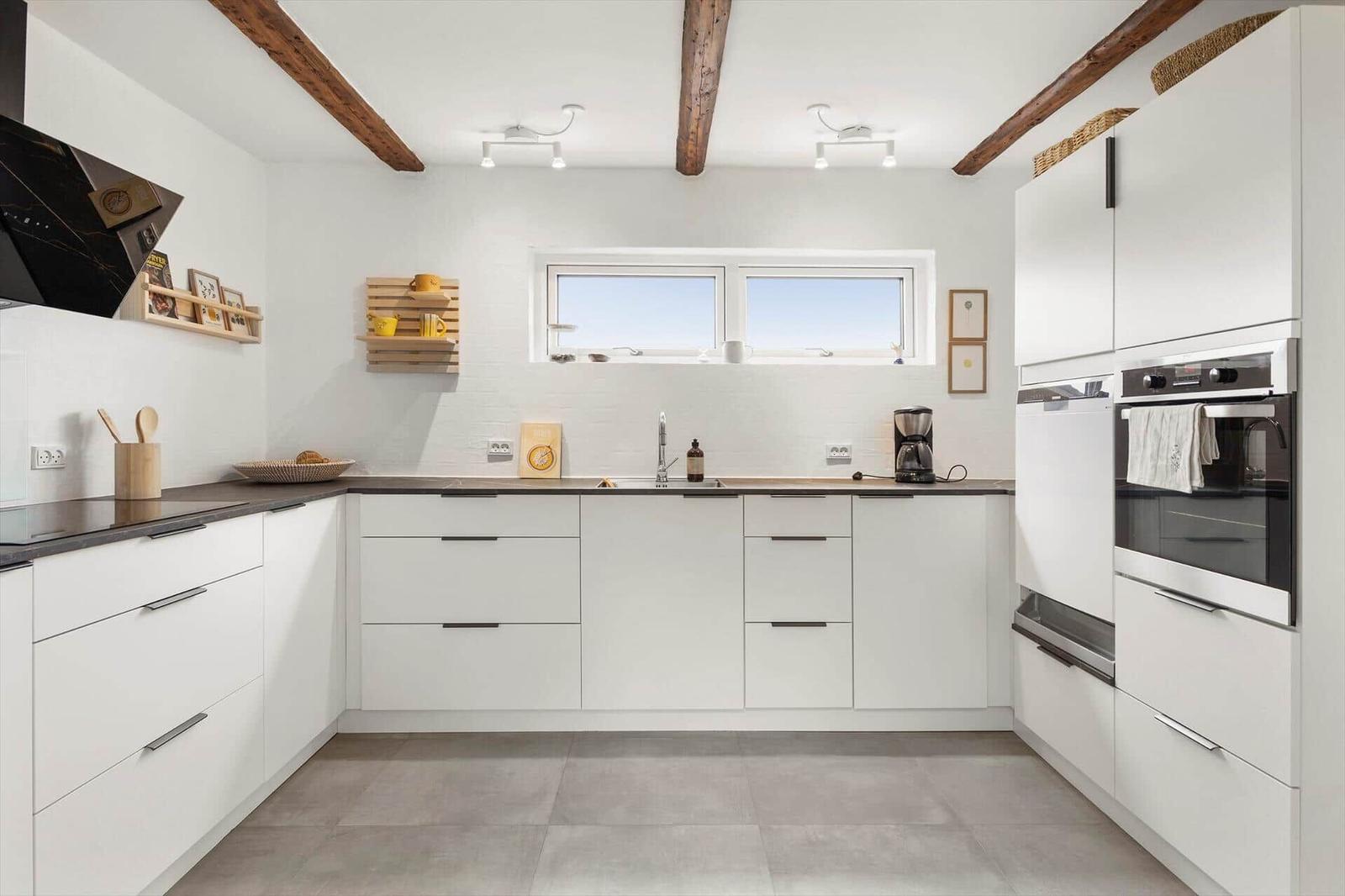 Moderne Küche mit weißen Schränken, dunkler Arbeitsplatte und Holzbalken an der Decke.