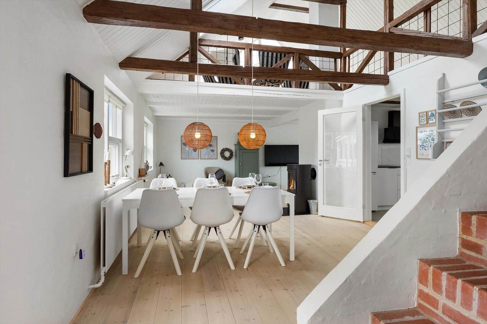 Essbereich mit Tisch, Stühlen und Holzbalken. Treppenhaus und offene Küche im Hintergrund.