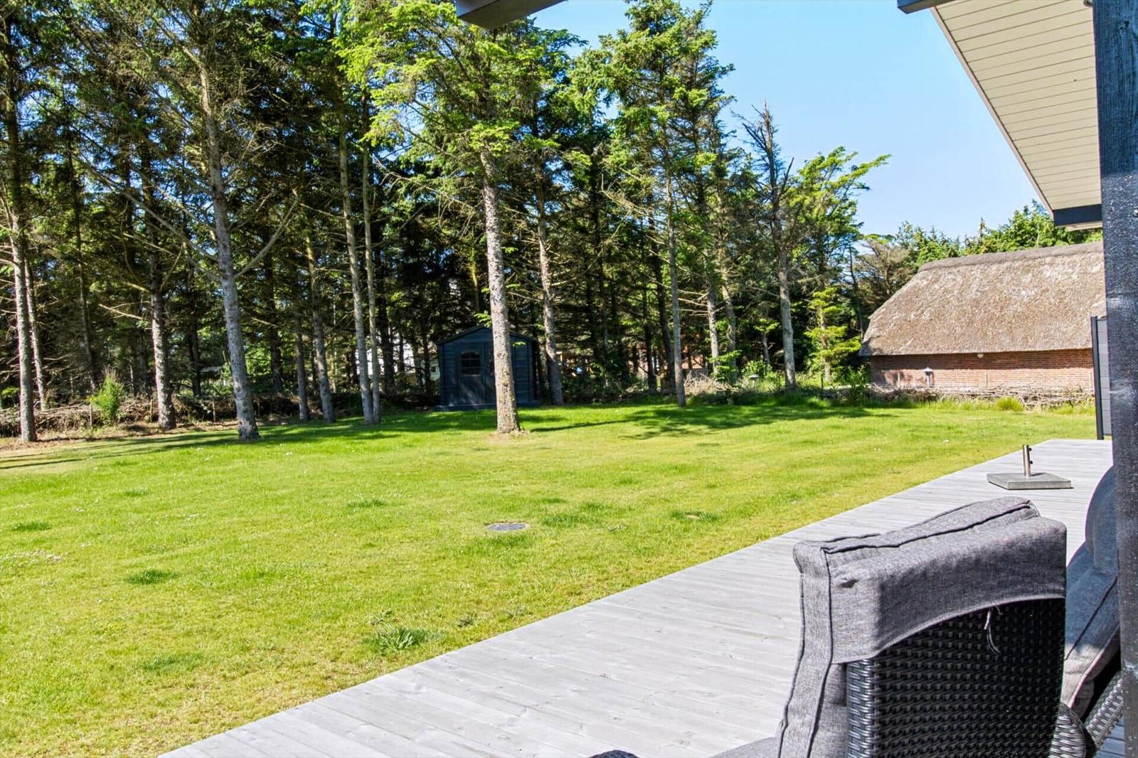 Holzterrasse mit Sitzmöbeln, grüner Rasen und Wald im Hintergrund.